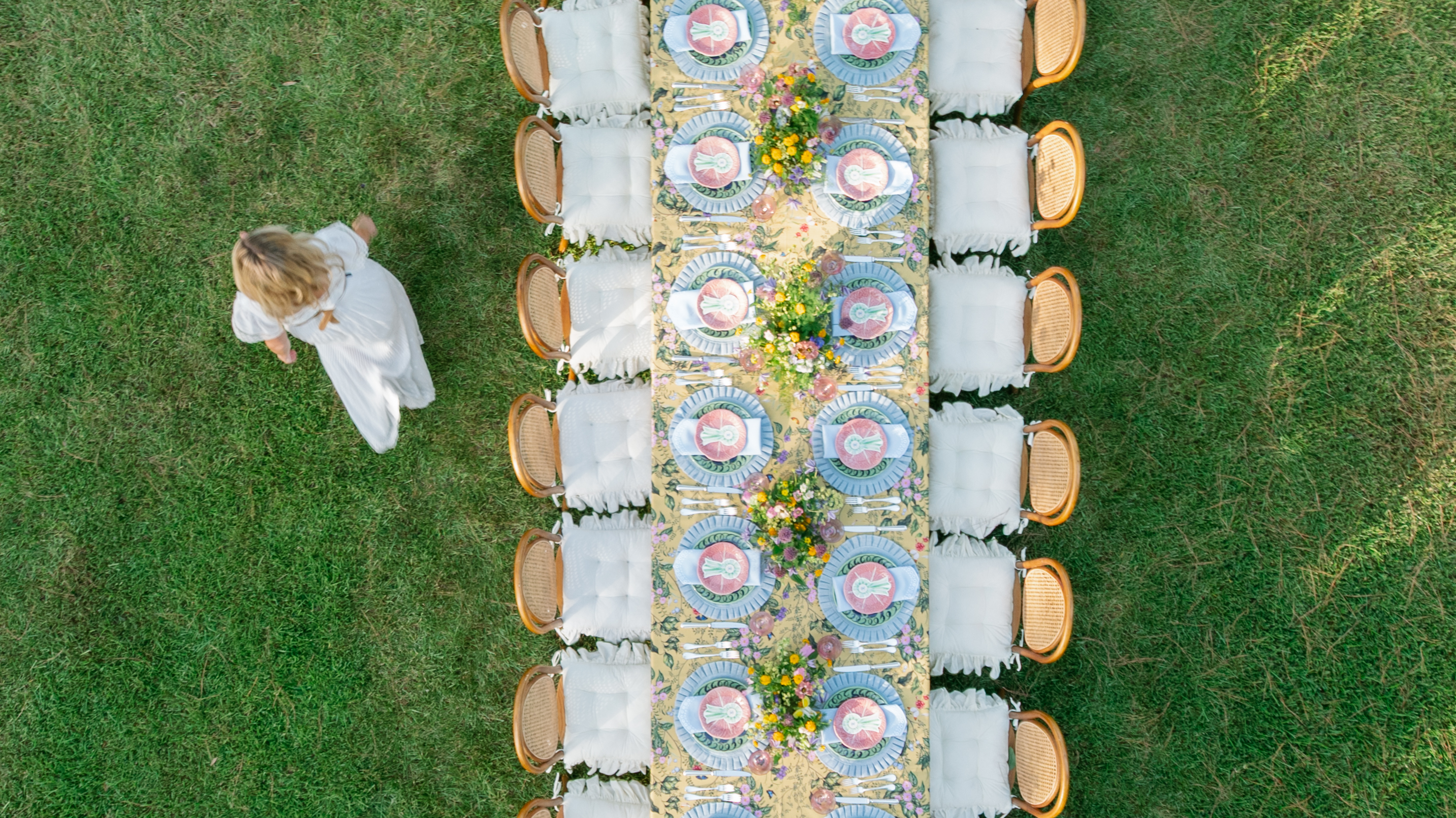 Overhead view of an outdoor dining table set on lawn with patterned yellow linens, colorful floral centerpieces, and cane chairs.