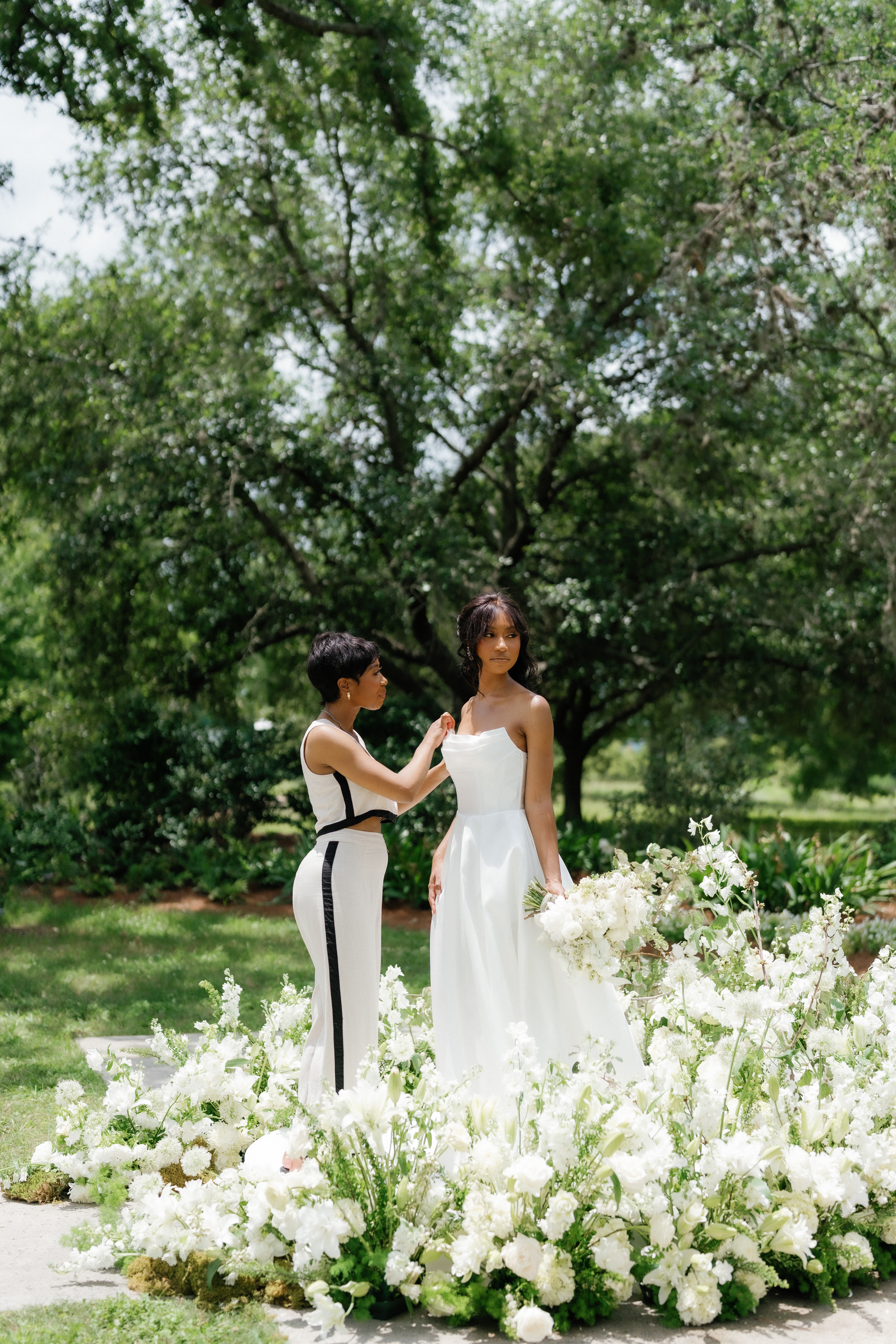 An attendant adjusts a bride's strapless white gown behind an abundant arrangement of white blossoms and greenery outdoors.