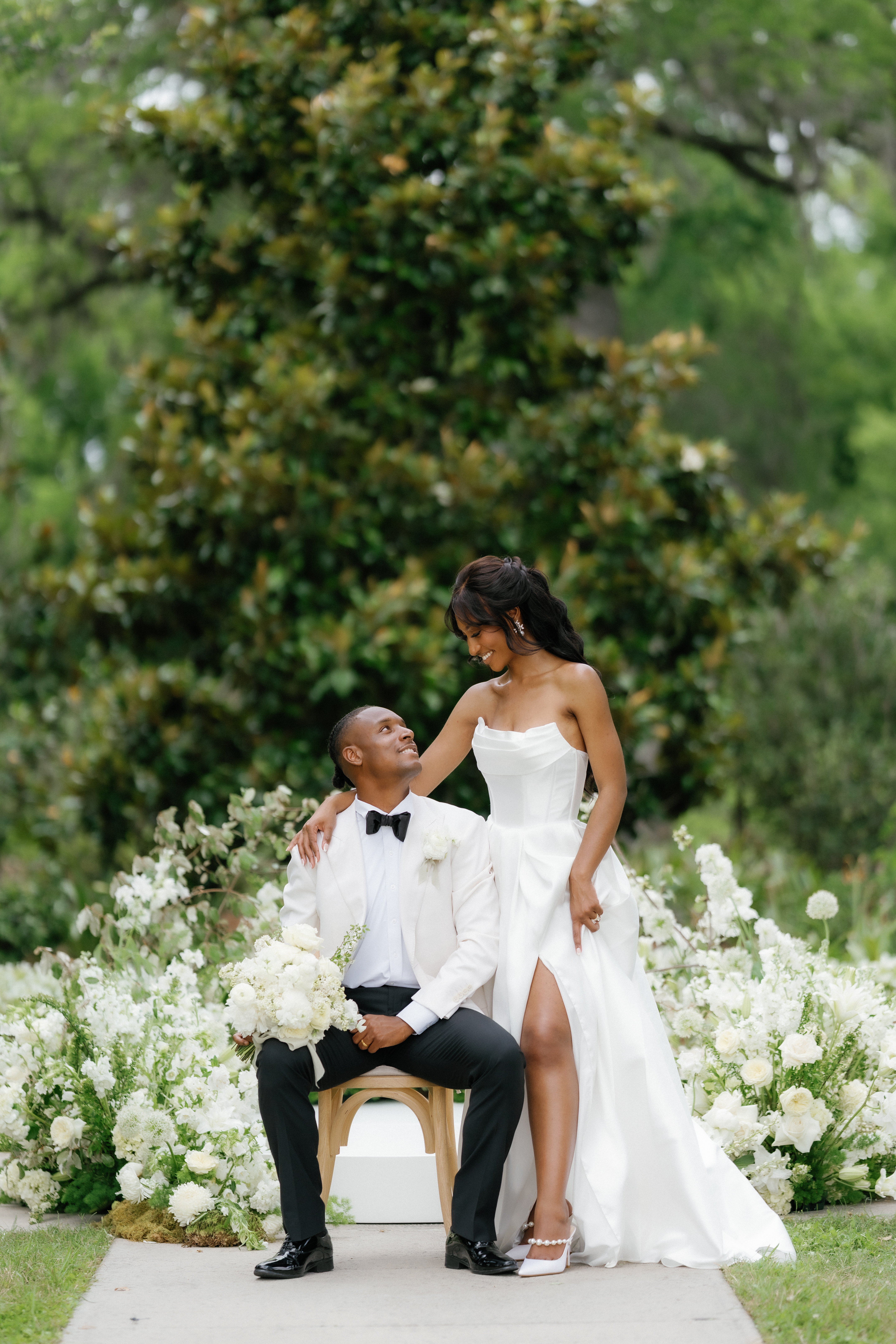 A bride in a strapless gown stands beside the seated groom in an ivory tuxedo against lush green foliage.