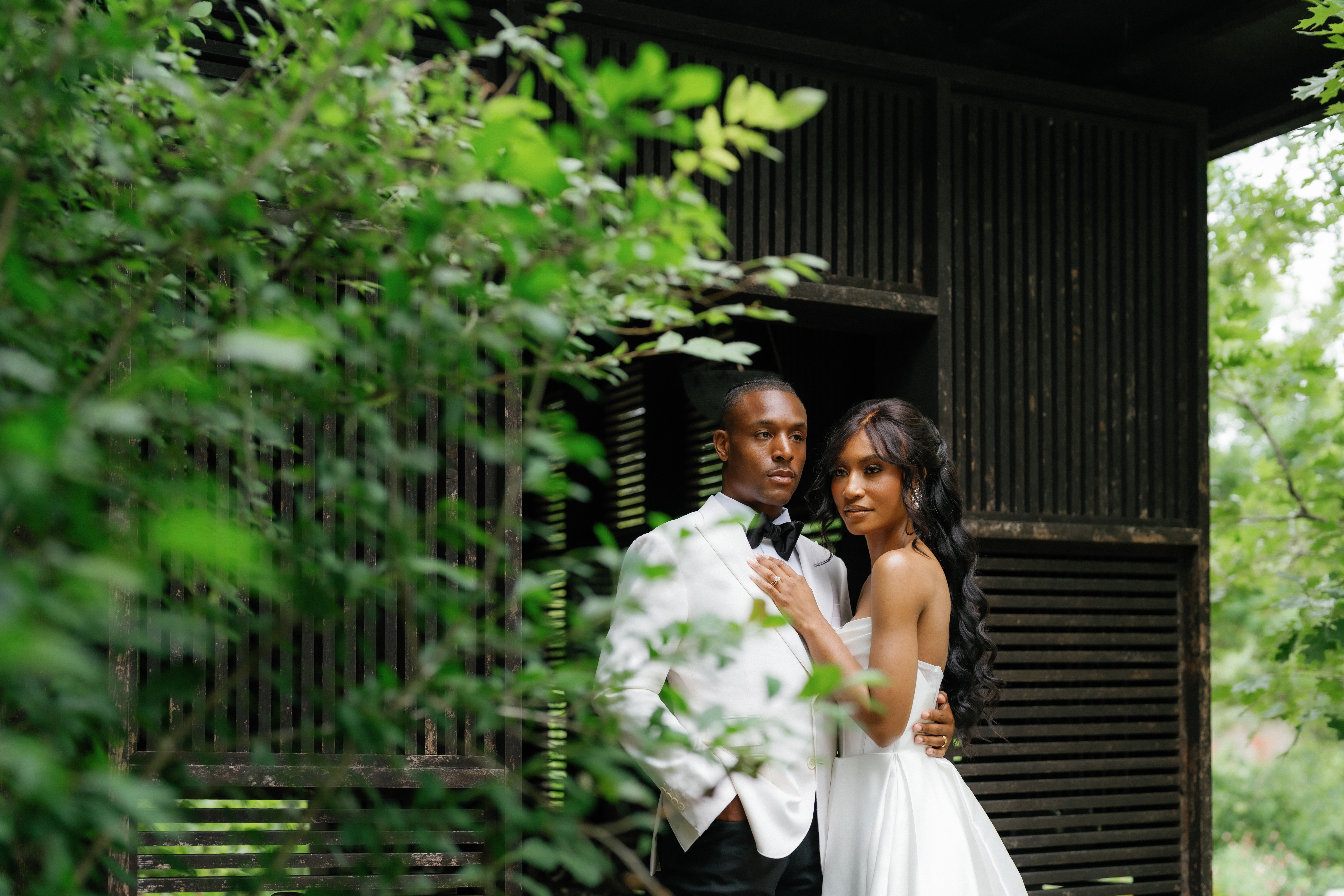 A groom in a white tuxedo and black bow tie stands amid lush green garden foliage as a hand rests gently on his lapel.