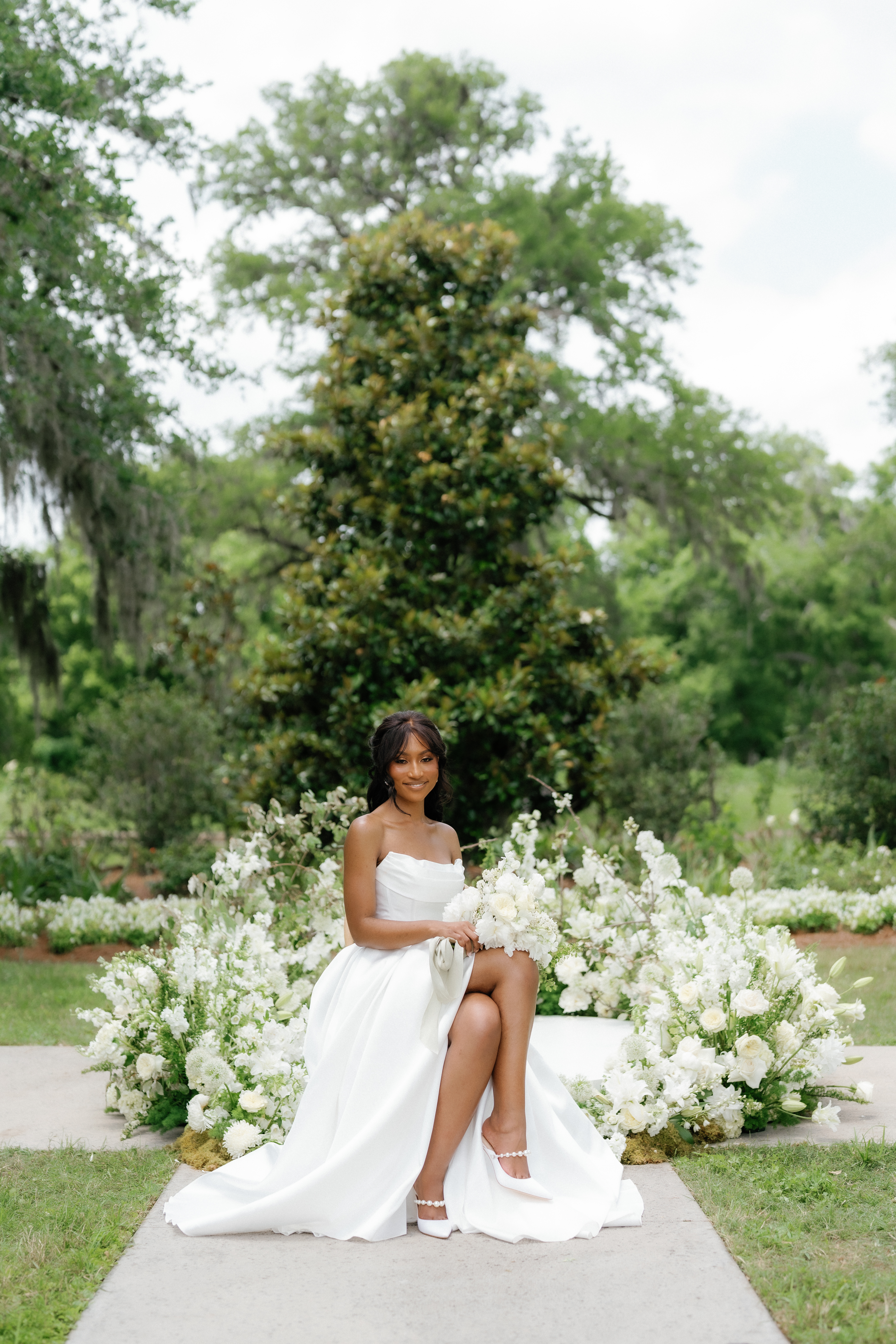 A smiling bride sits outdoors in a strapless white gown, holding a lush white bouquet against leafy green garden trees.