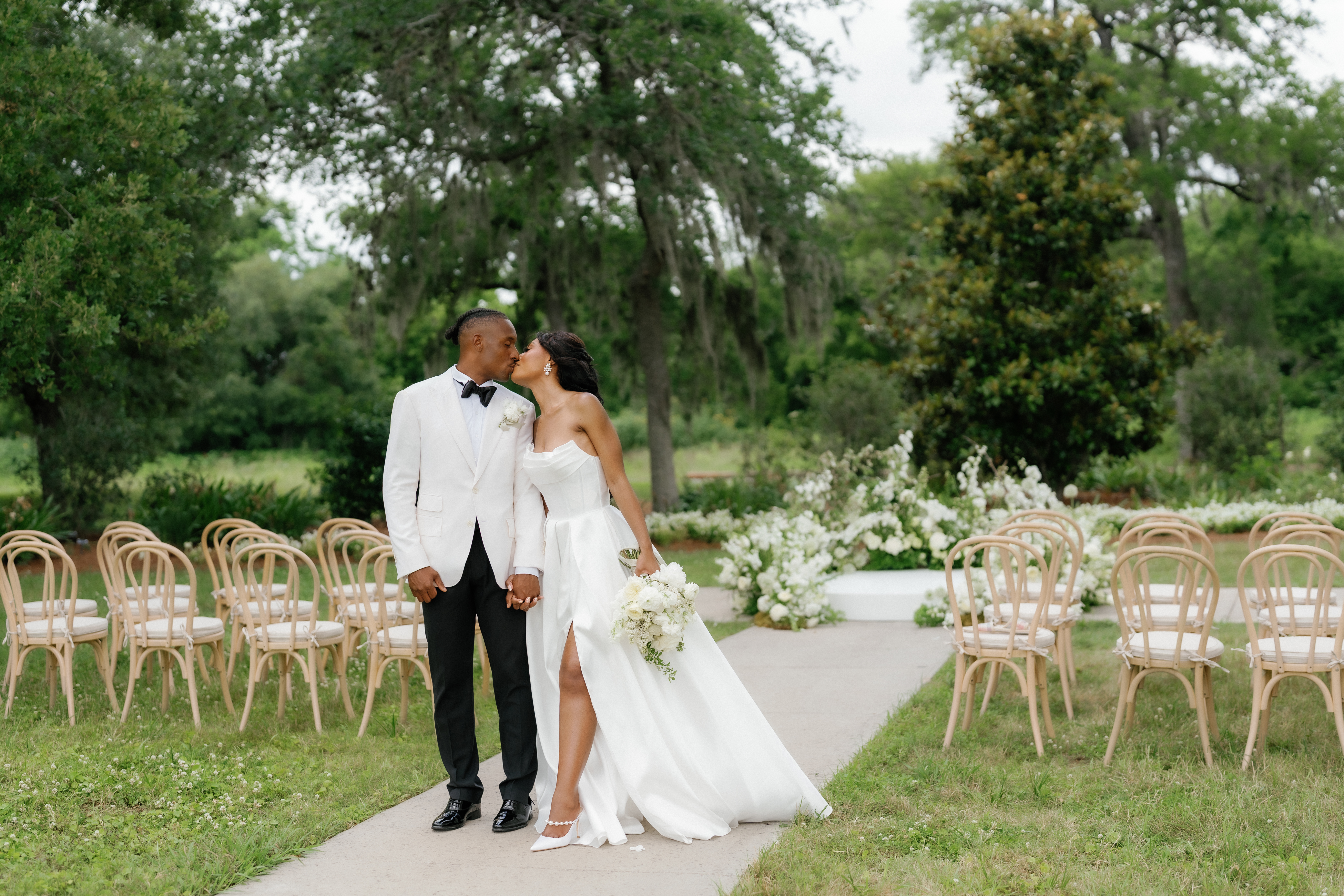 A bride in a white gown holds an all-white bridal bouquet while walking along an outdoor garden path lined with floral arrangements.