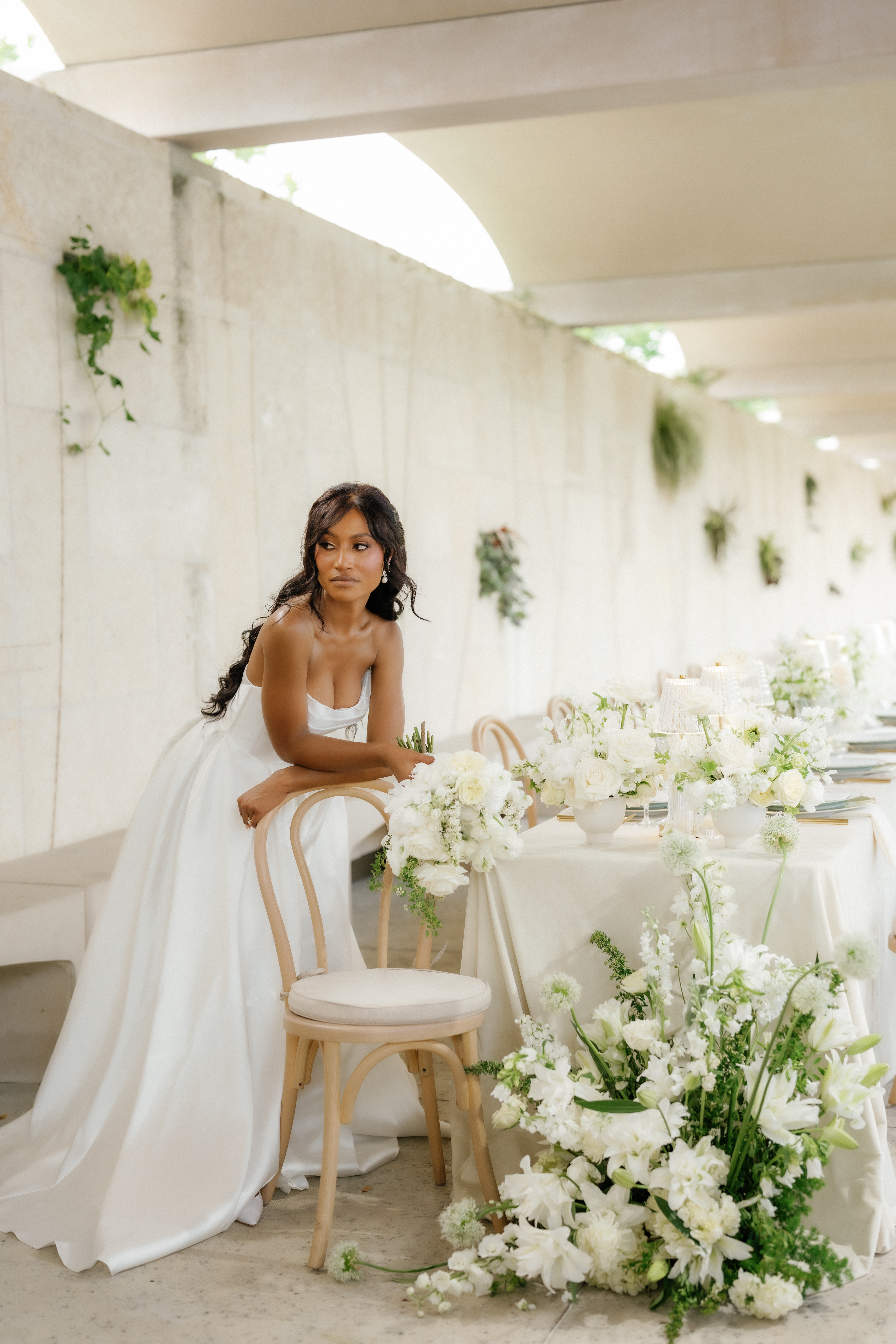 A bride sits at a reception table decorated with lush white floral arrangements and light wood chairs.