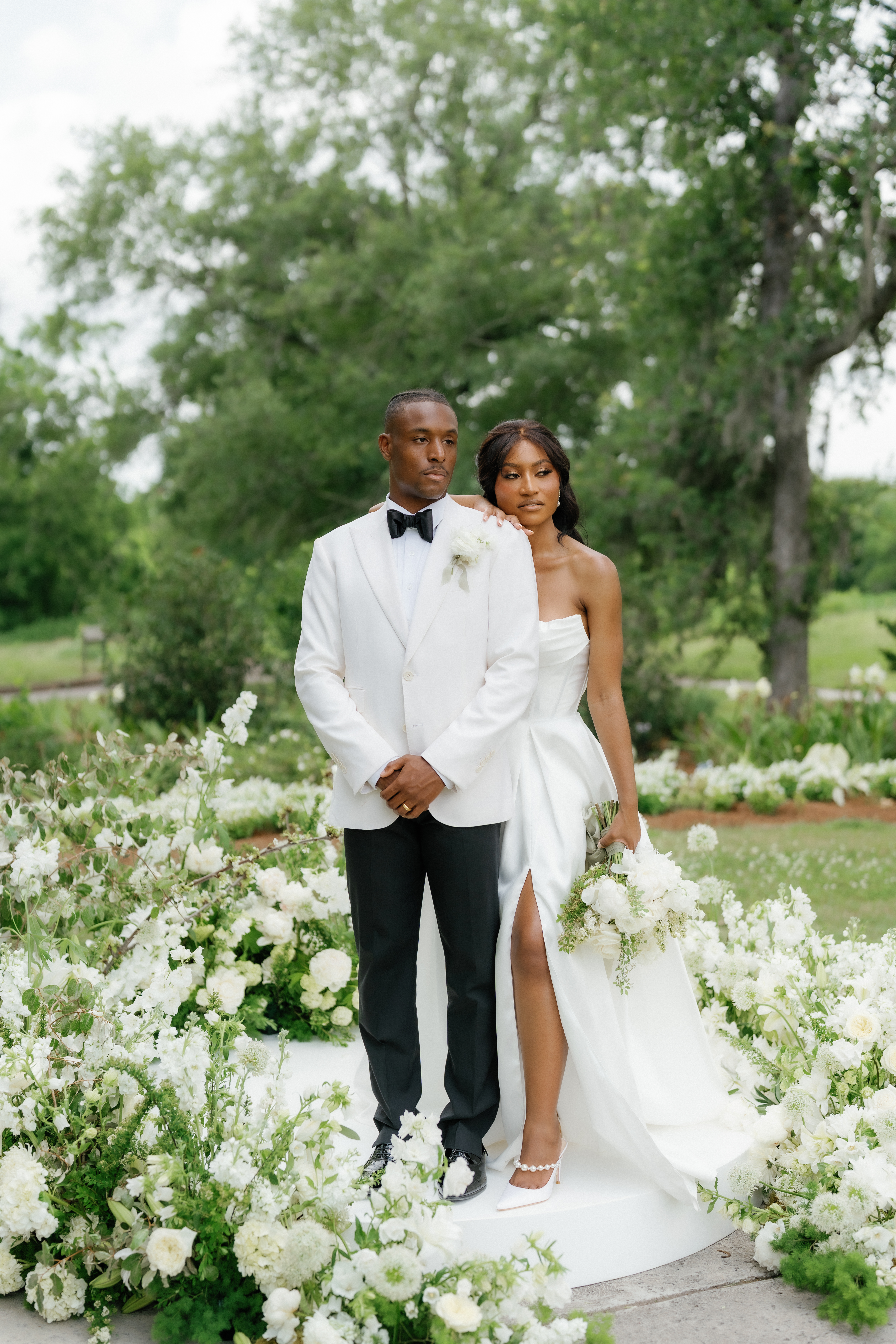 A bride in an ivory gown poses beside her groom in a white tuxedo jacket alongside lush white floral arrangements outdoors.
