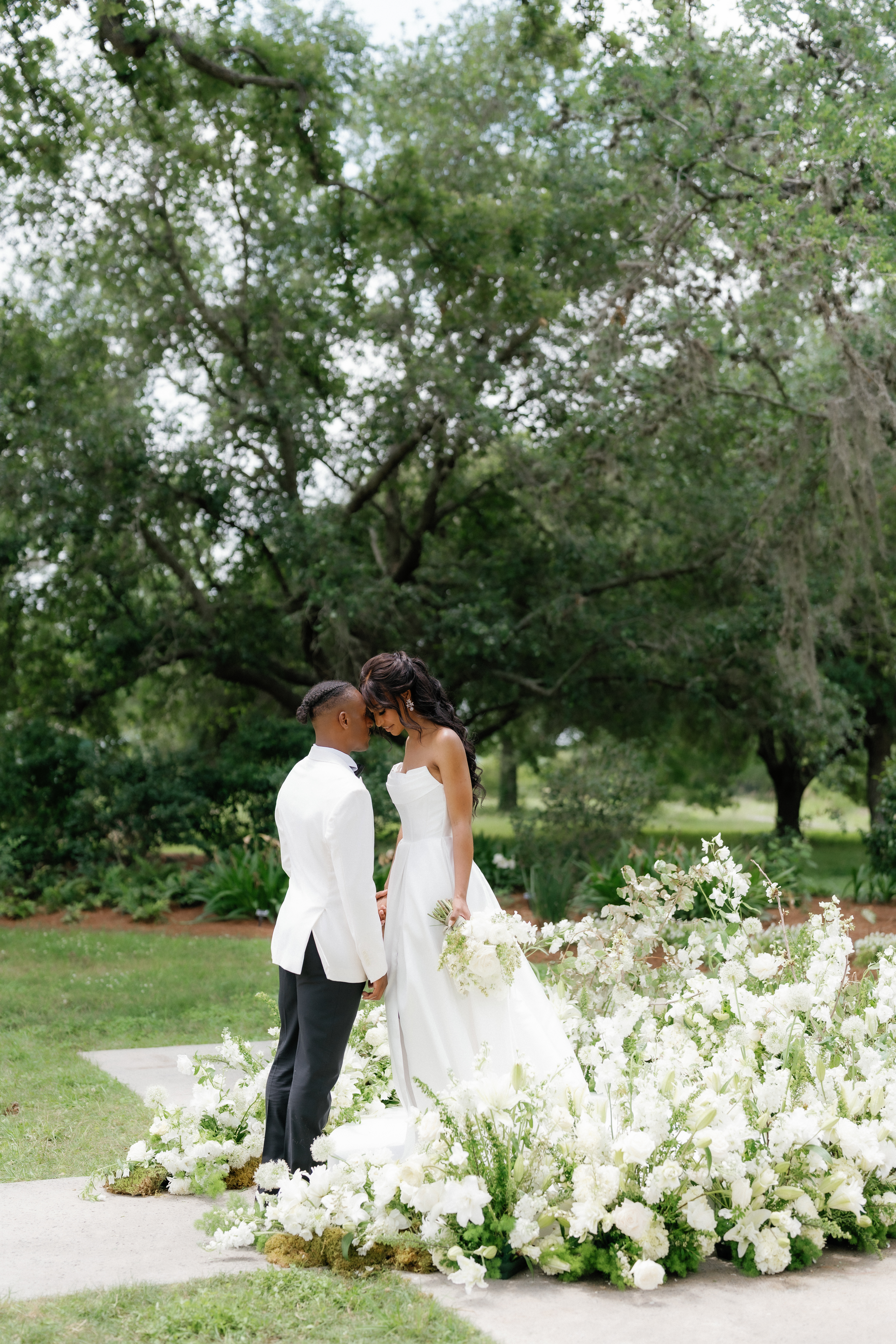 A bride and groom share a quiet moment surrounded by lush white floral arrangements along an outdoor garden path.