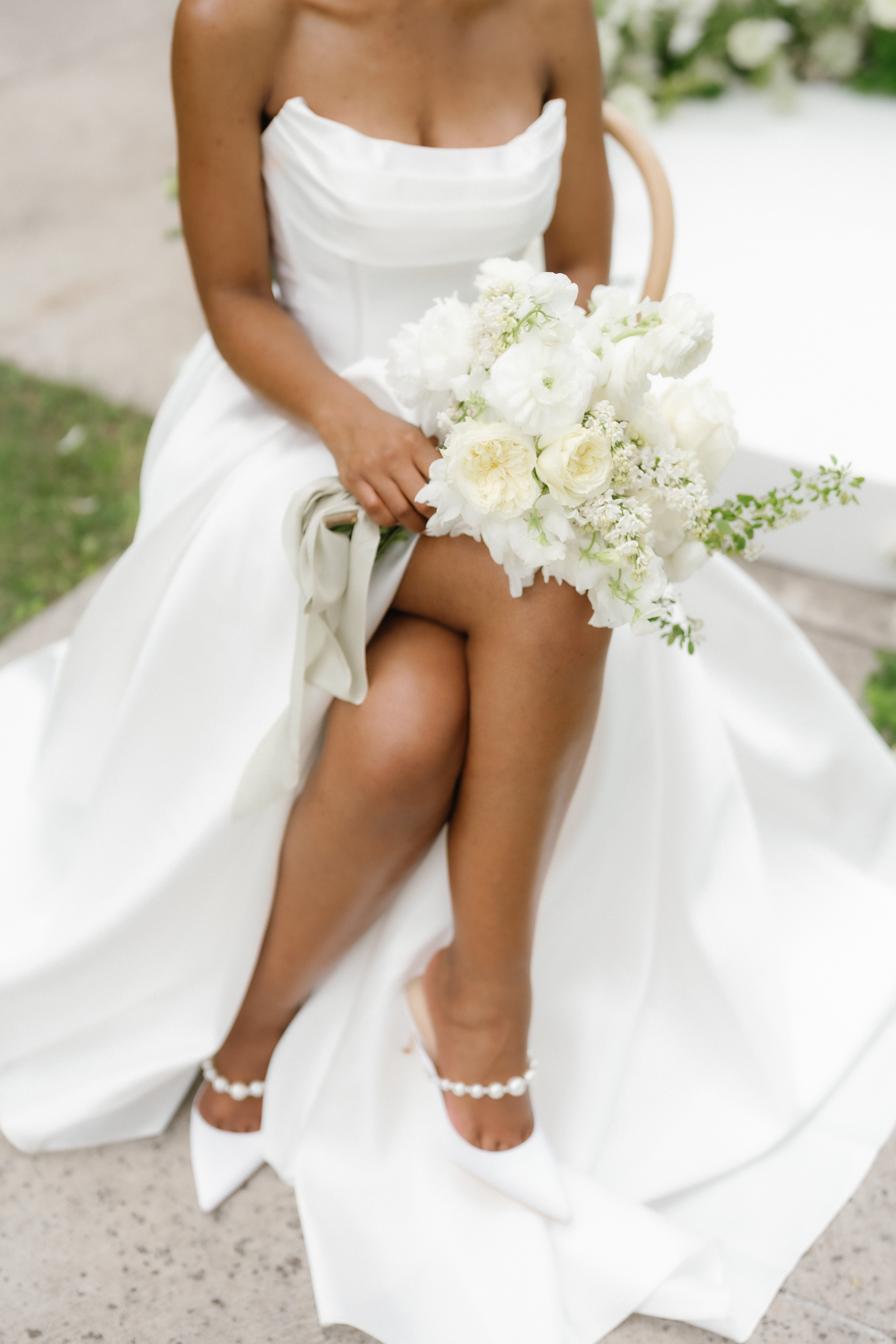 A seated bride in a strapless gown holds a monochromatic bouquet of creamy garden roses and delicate white ranunculus.