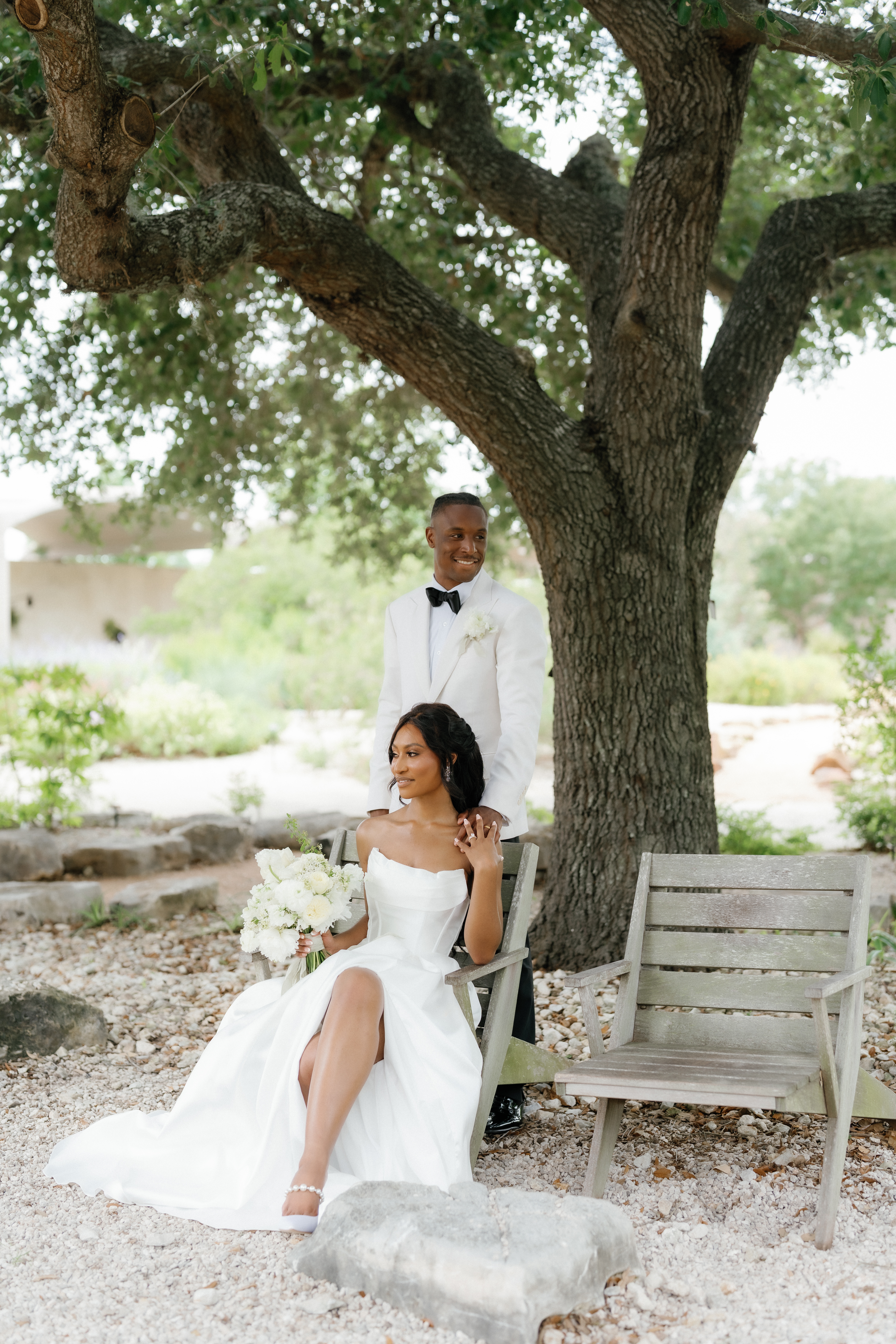 A bride in a strapless white gown sits on a bench beneath a large tree as the groom in an ivory tuxedo stands behind her.