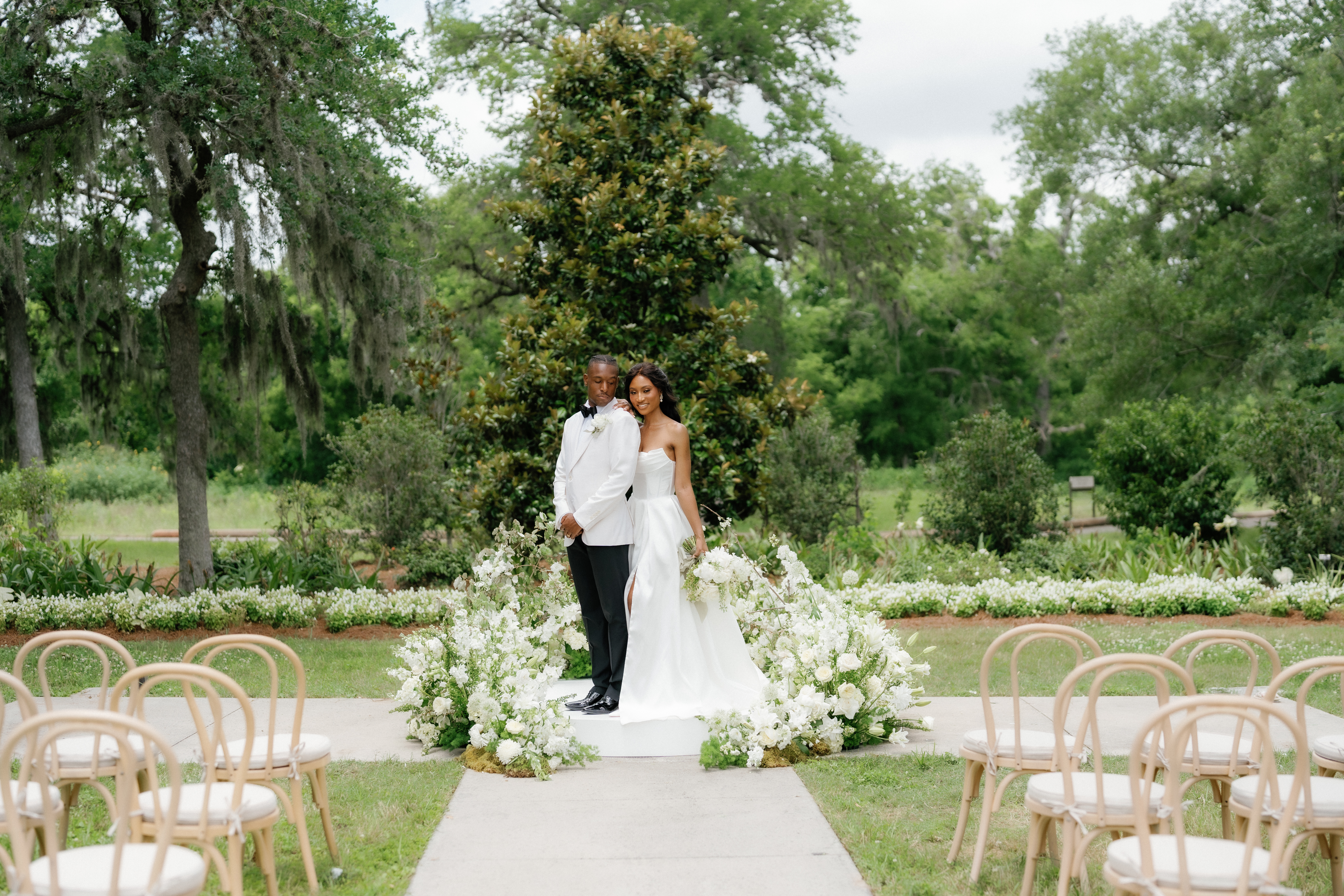 A bride in a strapless gown and groom in a white tuxedo stand together on an outdoor aisle framed by white florals and lush trees.