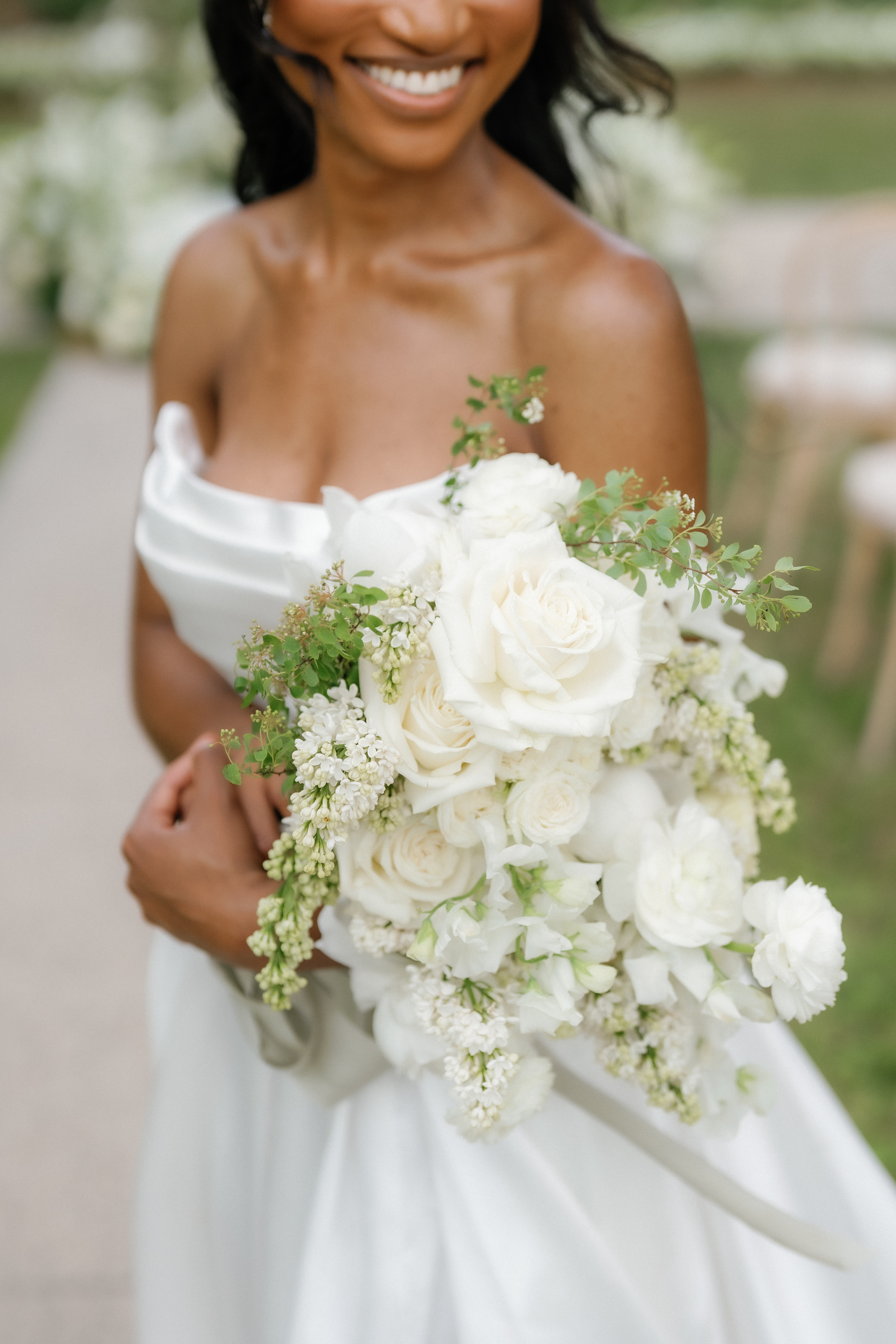 A smiling bride holds a lush bridal bouquet of ivory roses, delicate white lilacs, and subtle greenery.