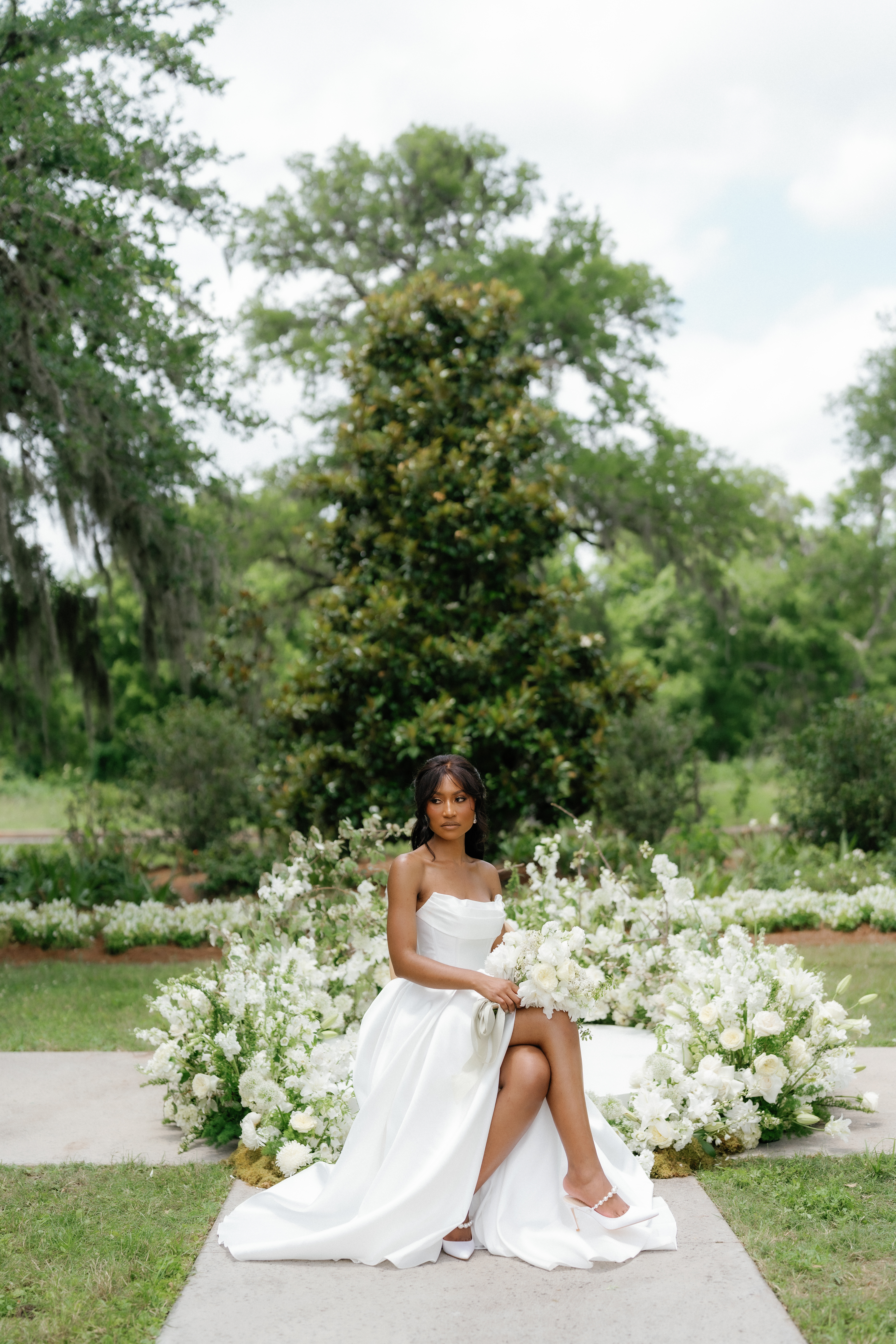 A seated bride in a strapless white gown holds a lush bouquet of ivory blooms in a lush garden setting.