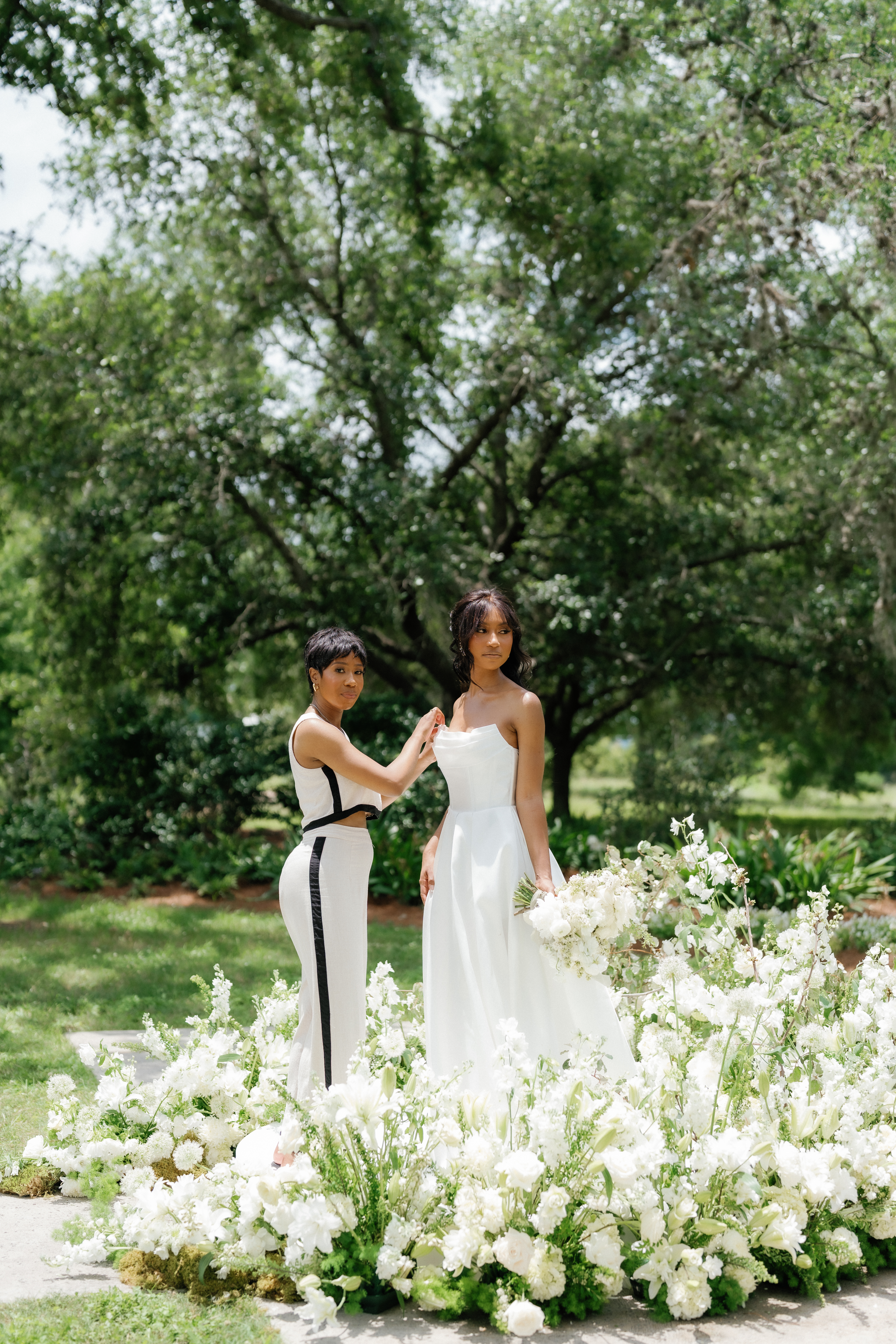 A bride in a strapless white gown stands beside lush white floor floral arrangements and garden greenery.