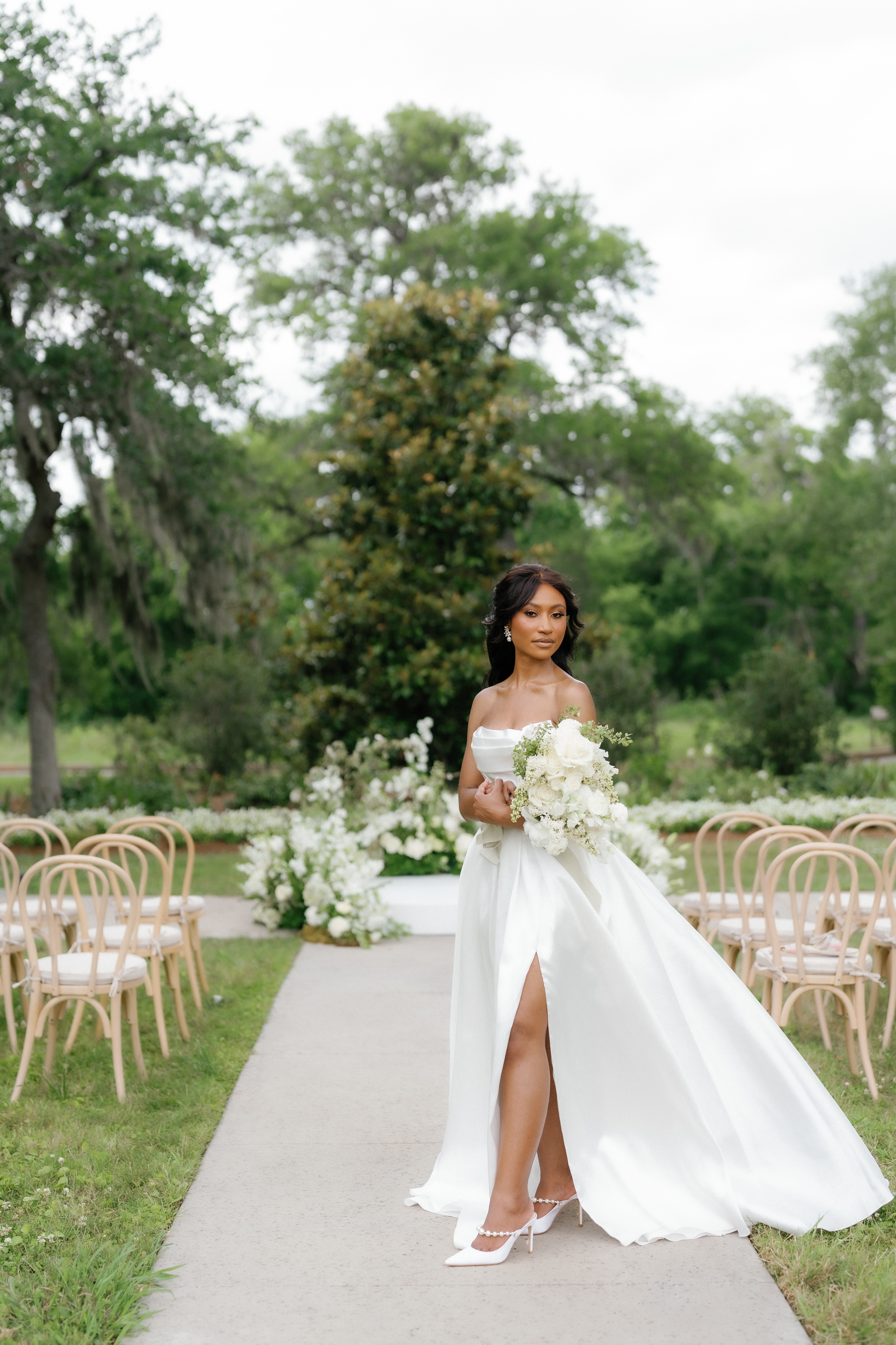 A bride in a strapless white gown holds a monochrome ivory bouquet outdoors against lush greenery.