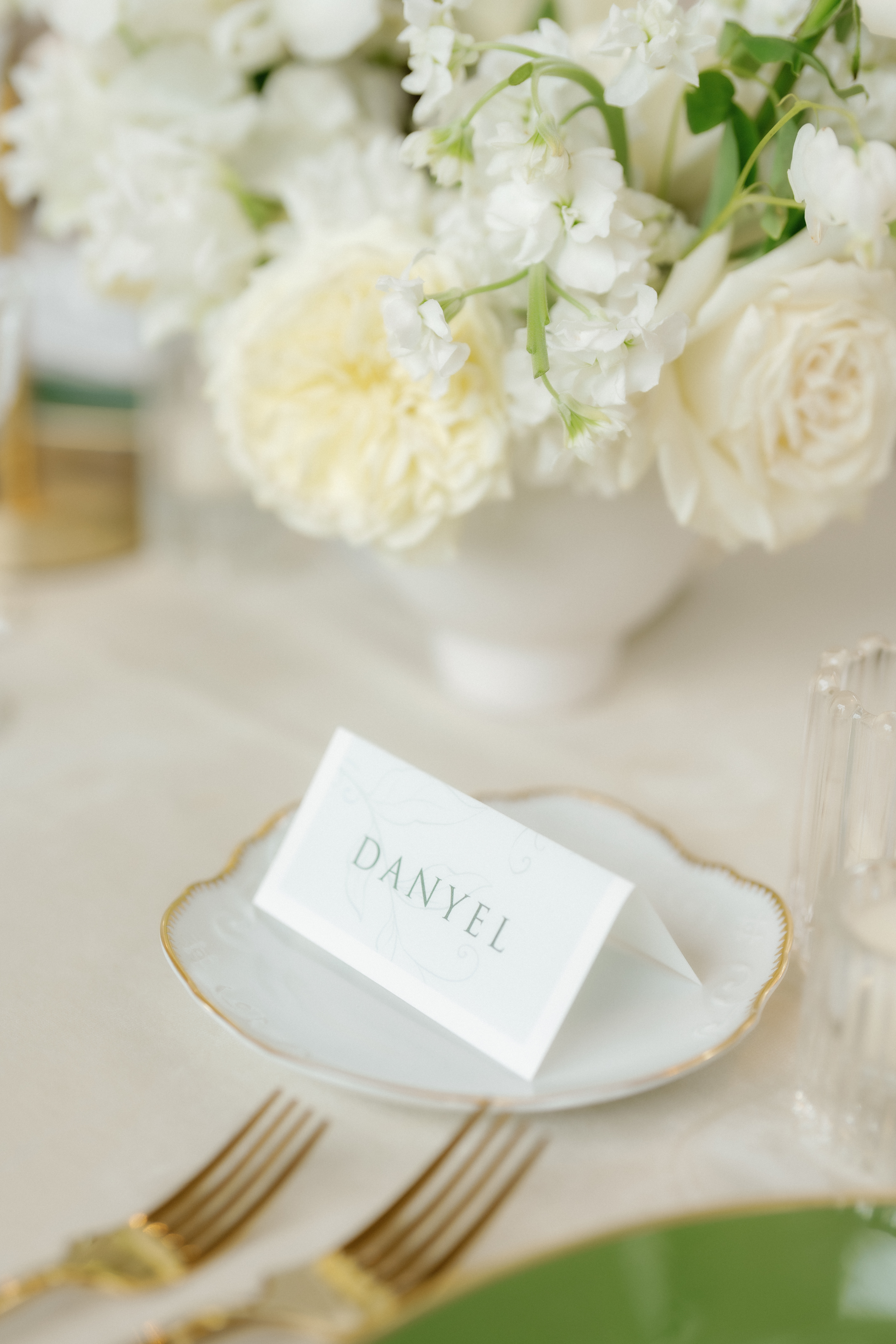 Soft white floral centerpiece beside a personalized place card and gold flatware at an elegant wedding table setting.