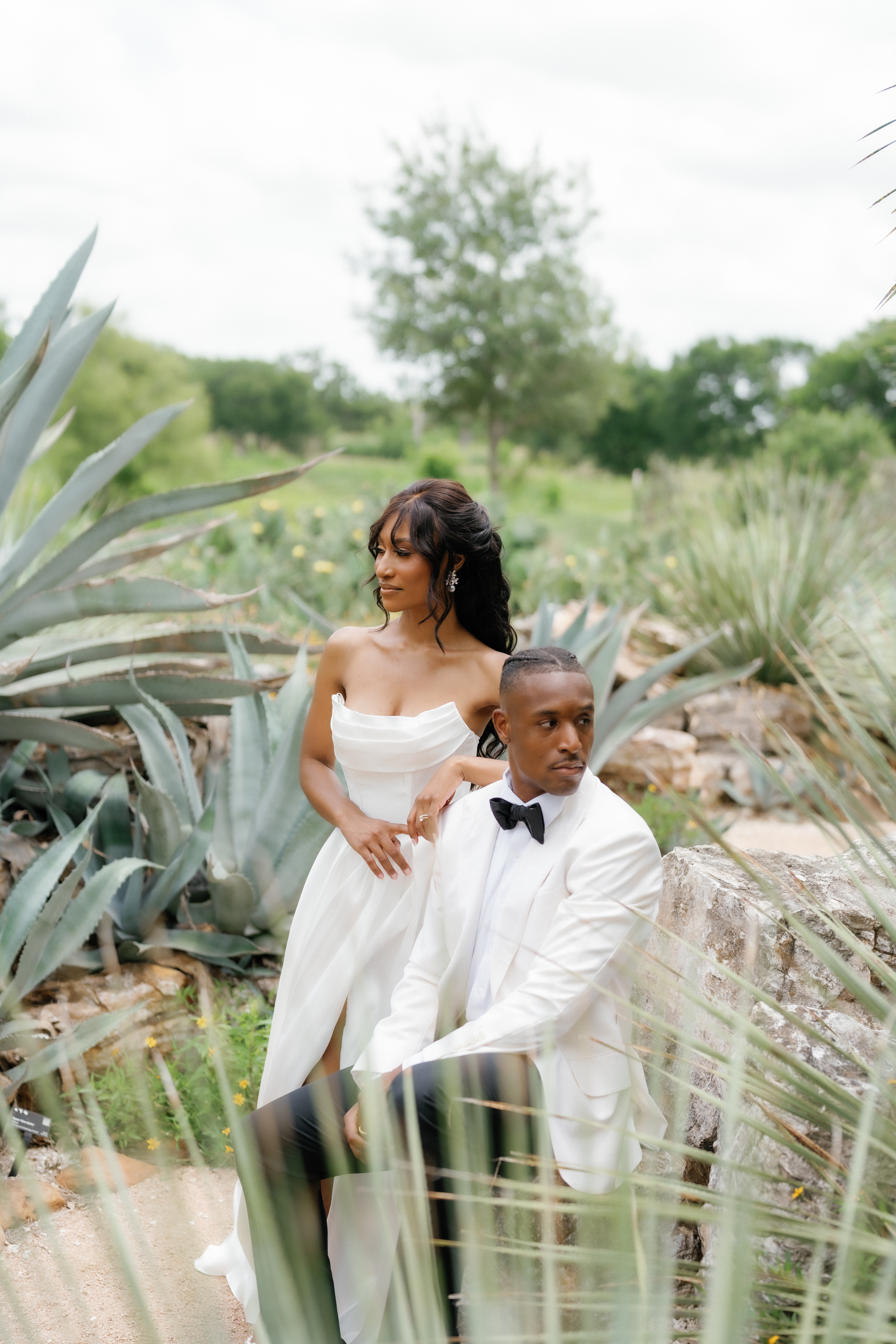 A bride in a strapless gown and a groom in an ivory tuxedo jacket pose together outdoors in a lush garden setting.