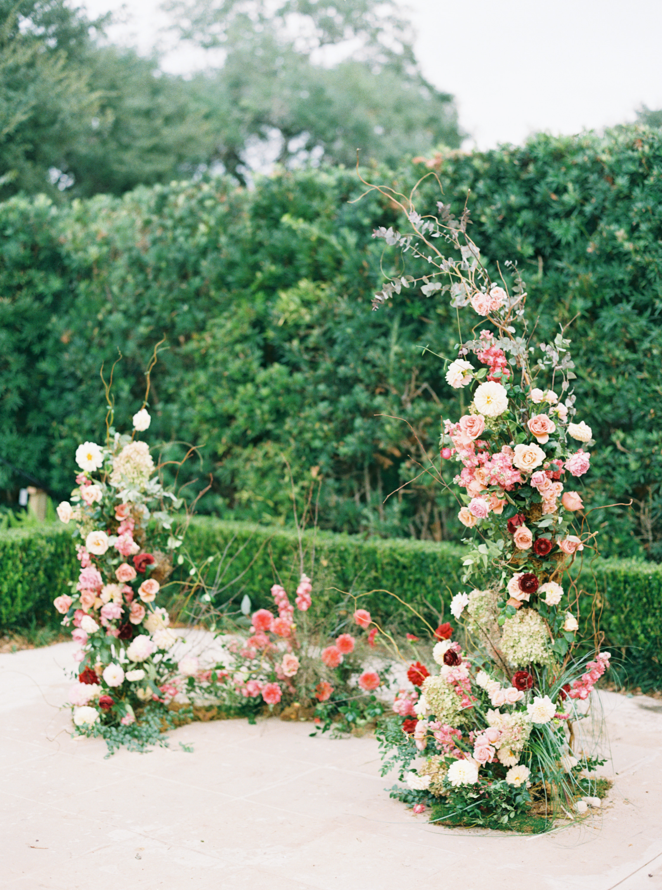 A romantic floral arch framed with pink roses, dahlias, and delicate branches lines a garden stone terrace.