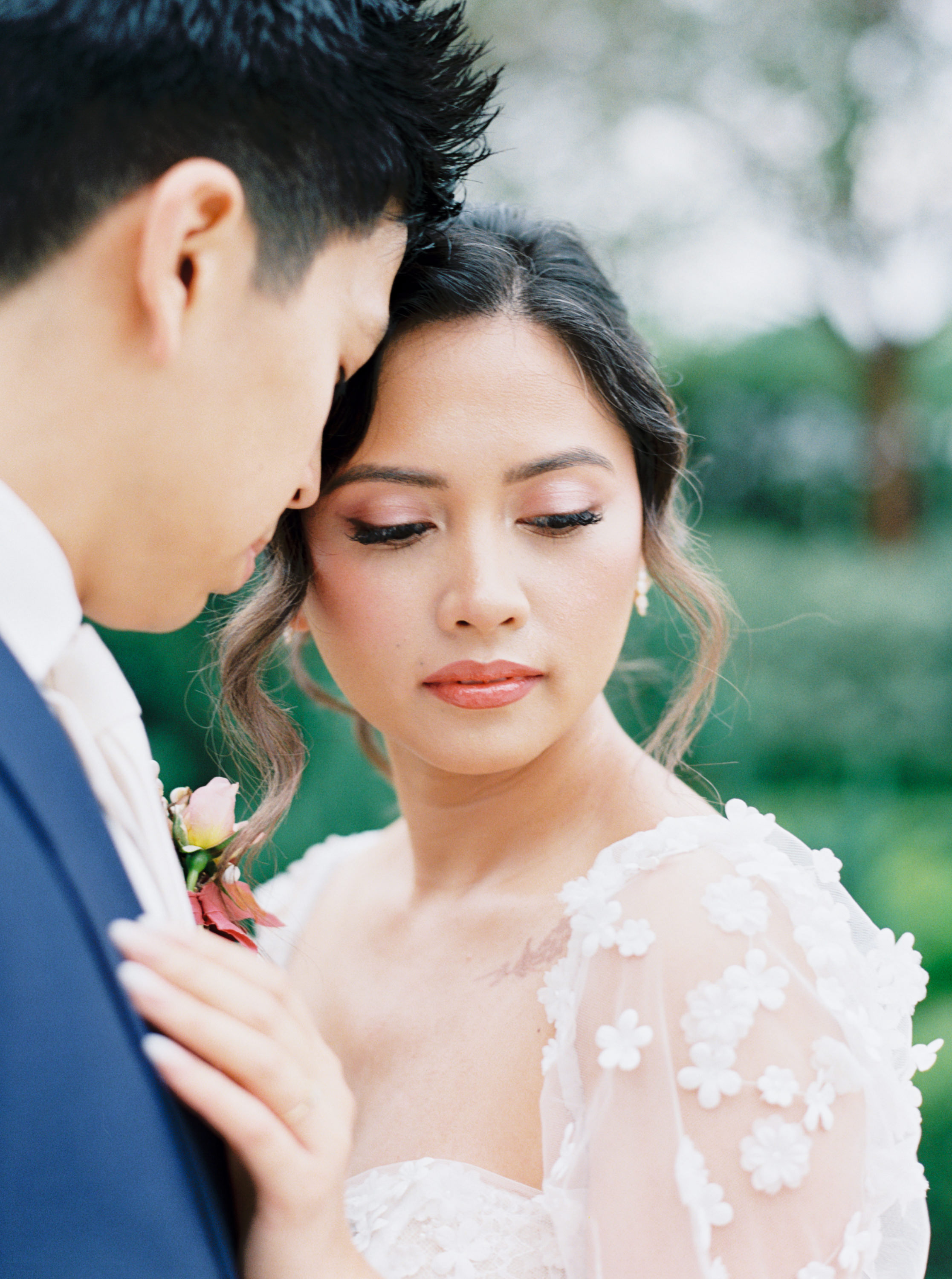 Close-up portrait of a bride wearing a floral appliqué wedding dress leaning softly against the groom outdoors.