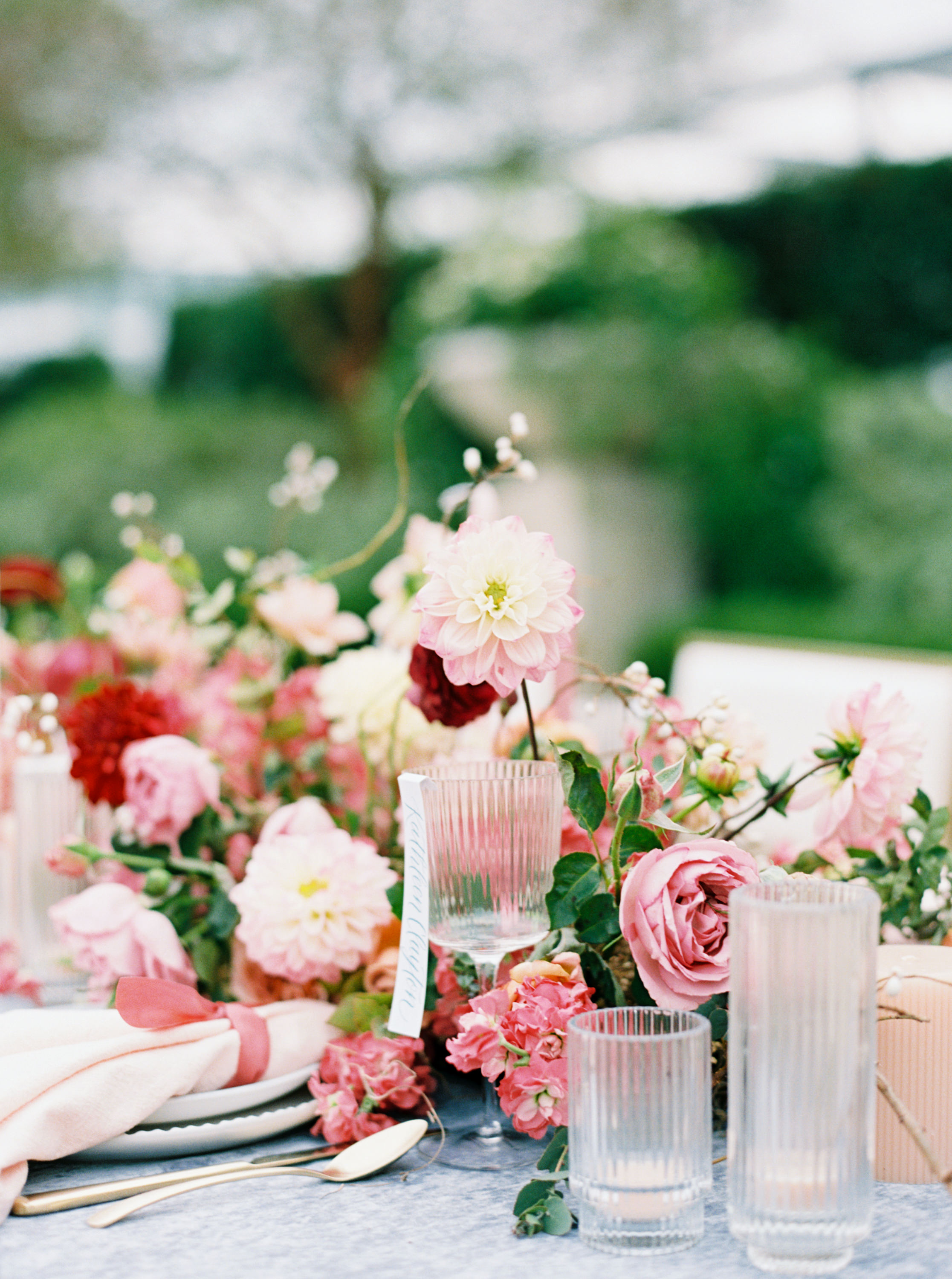 Pink dahlias and garden roses accent an elegant outdoor reception table set with fluted glassware and gold cutlery.