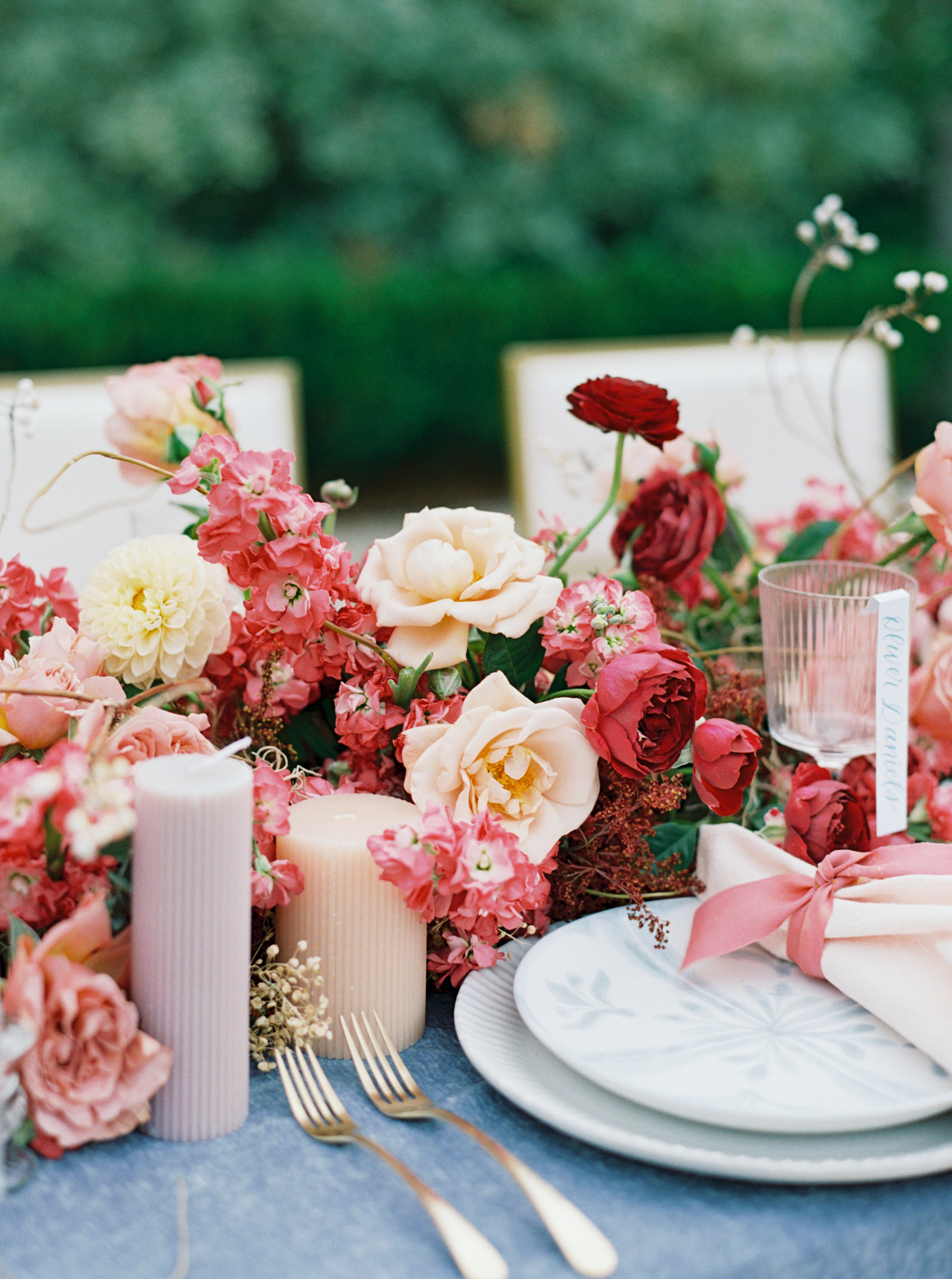 Blush and deep red roses with pink florals accent a wedding table setting with ribbed candles and gold flatware.