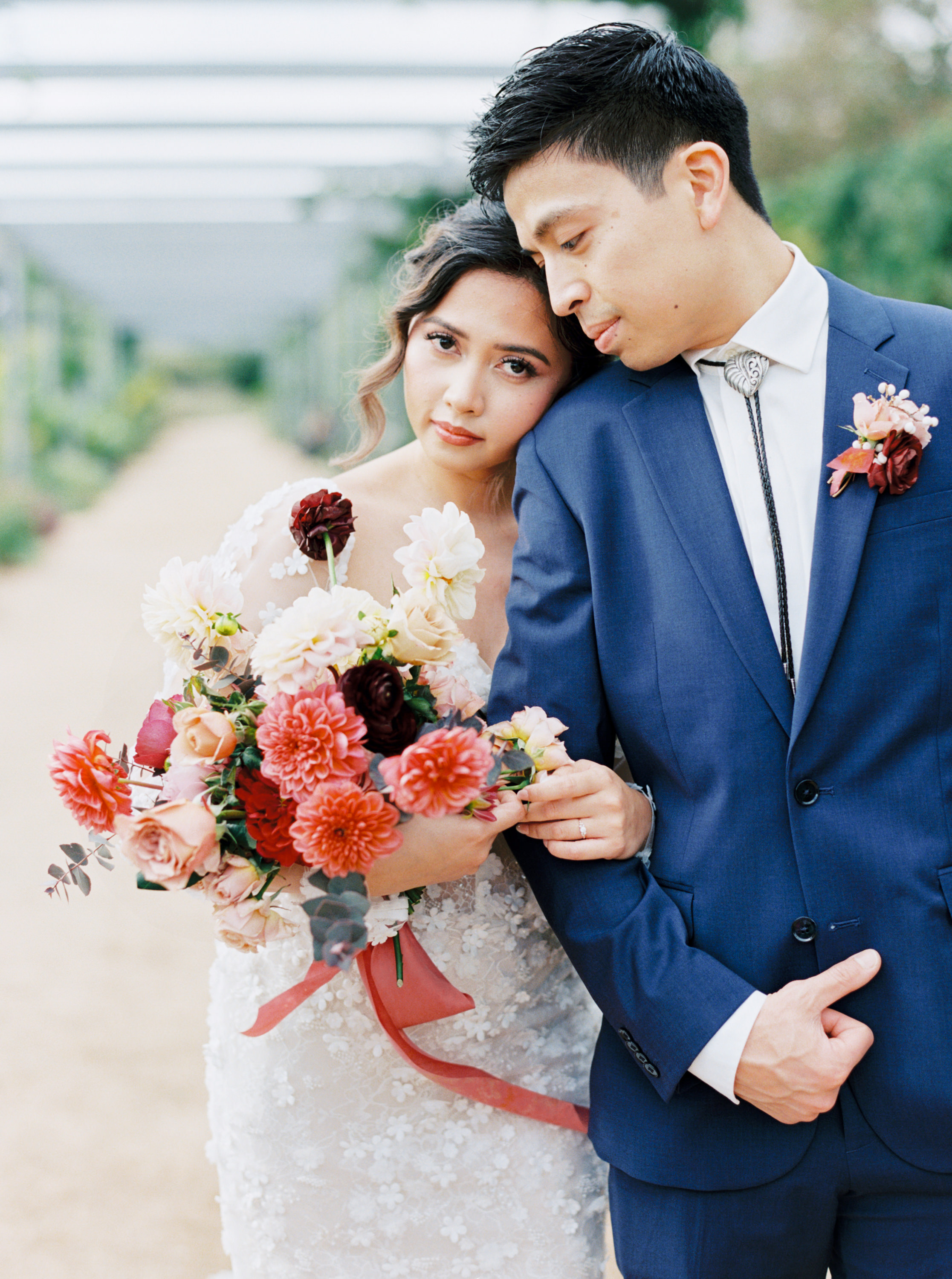A bride holds a vibrant coral, blush, and burgundy bouquet while leaning against the groom in a navy suit.