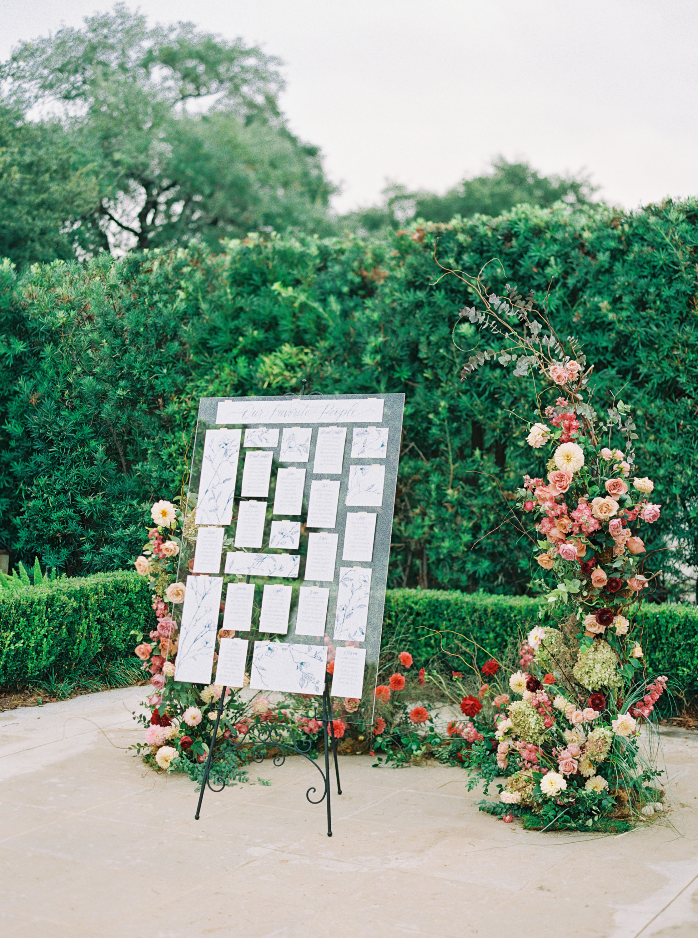 An acrylic seating chart on a wrought iron easel framed by lush blush, coral, and red garden flowers against green hedges.