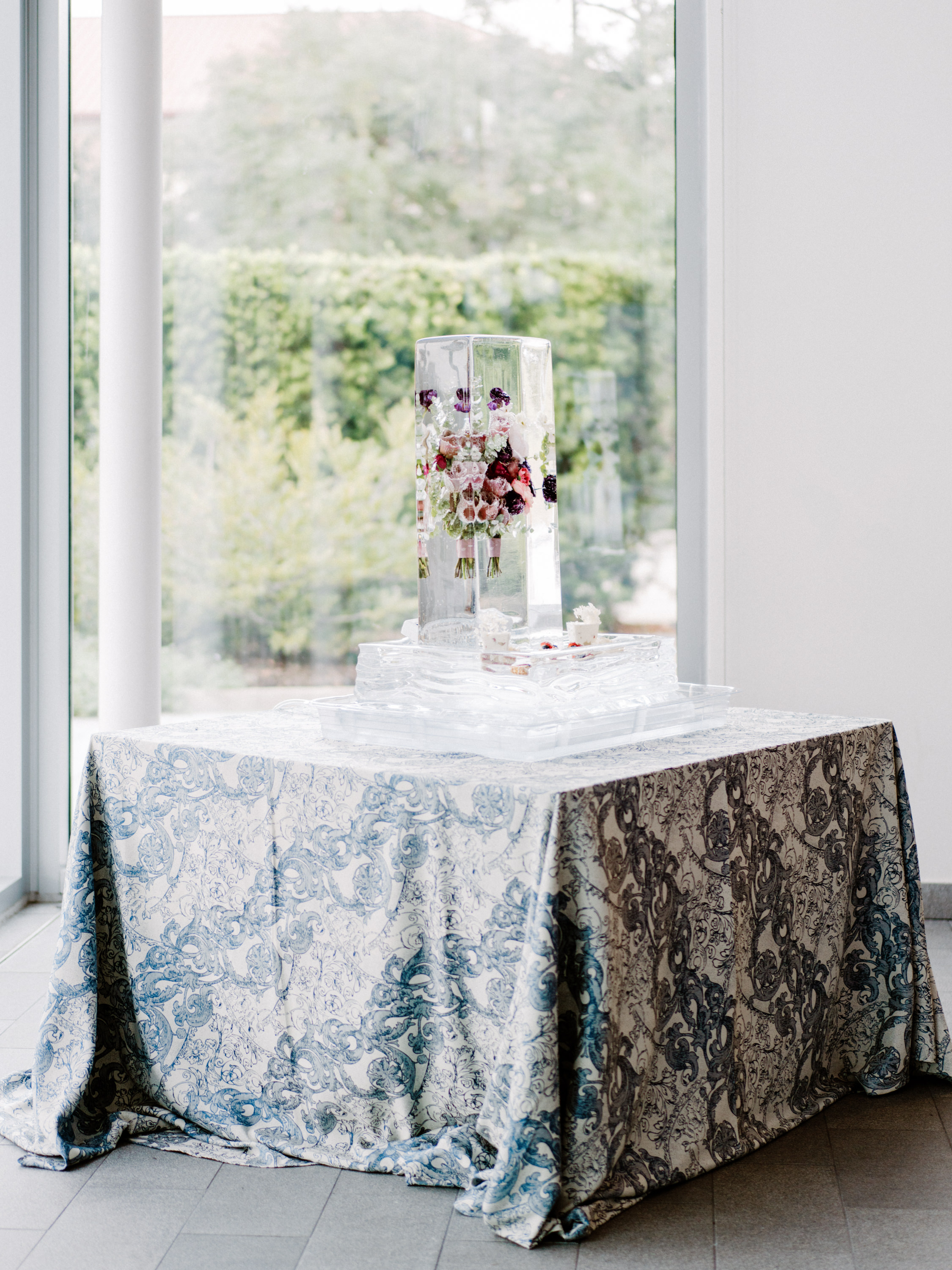 A tall clear ice sculpture encasing suspended floral bouquets sits on a blue and white patterned table.