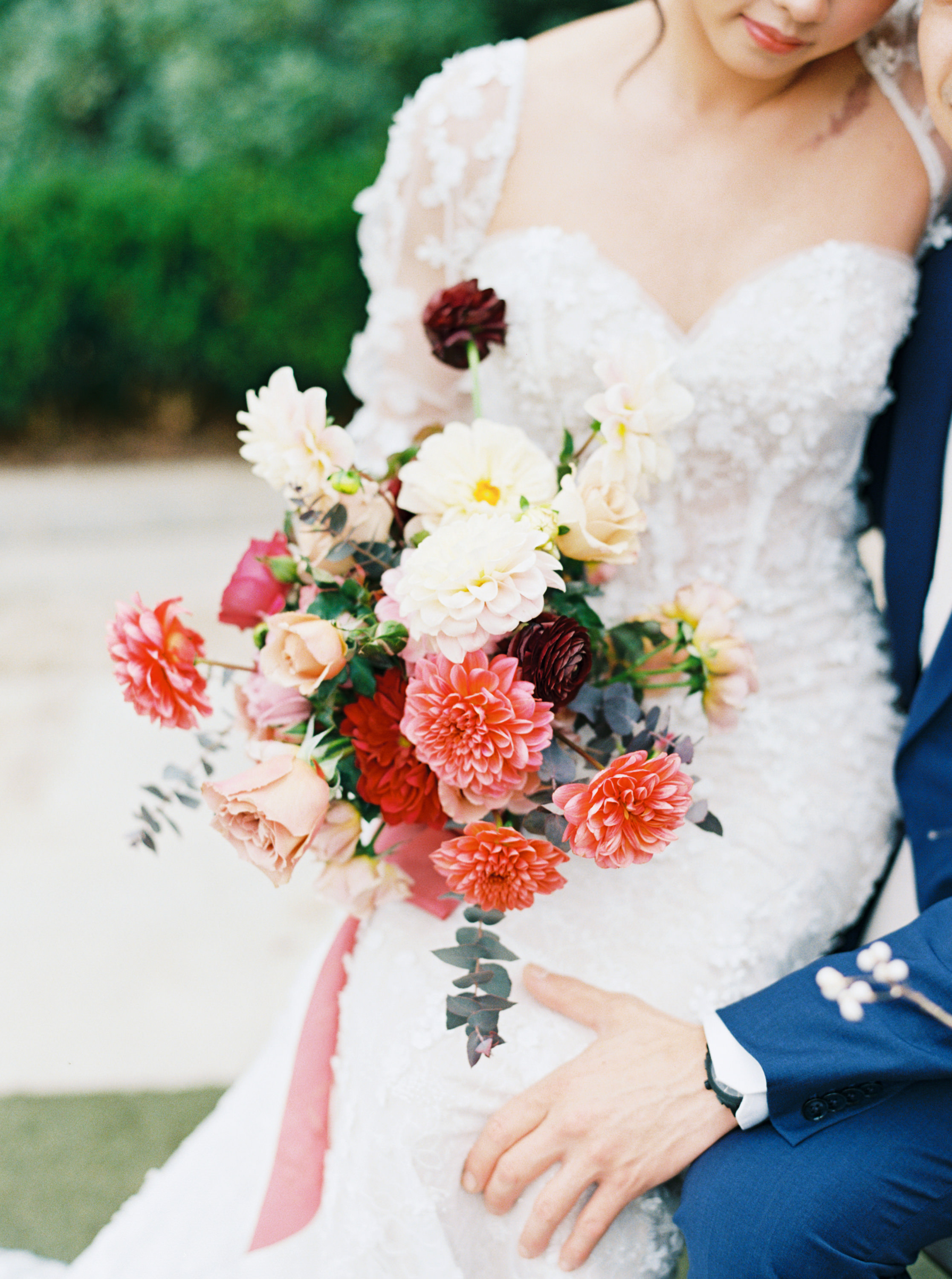 A bridal bouquet of coral dahlias, blush garden roses, and deep burgundy blooms against an intricately beaded lace gown.