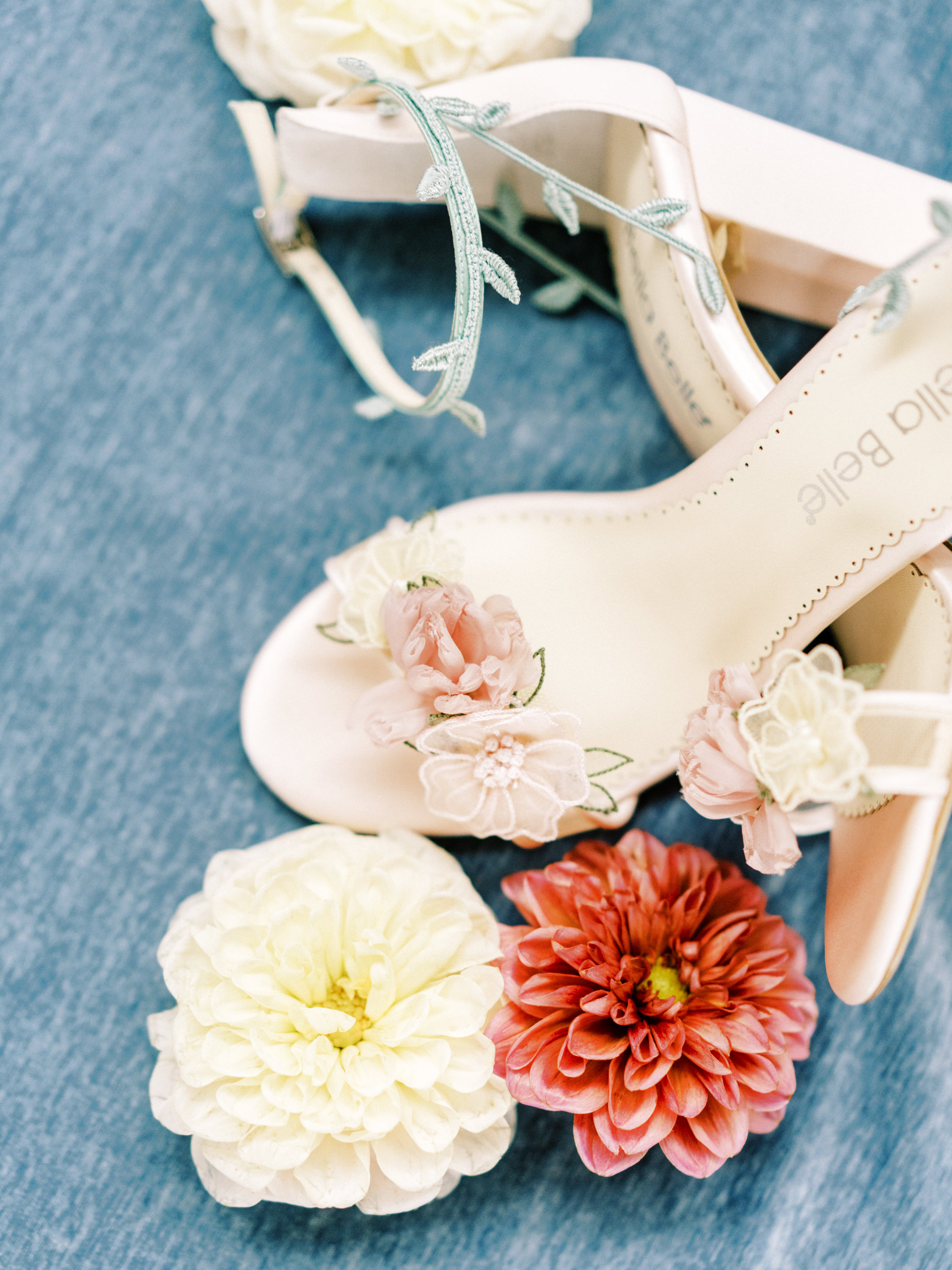 Embellished bridal heel styled with fresh cream and coral dahlias on a textured blue surface.