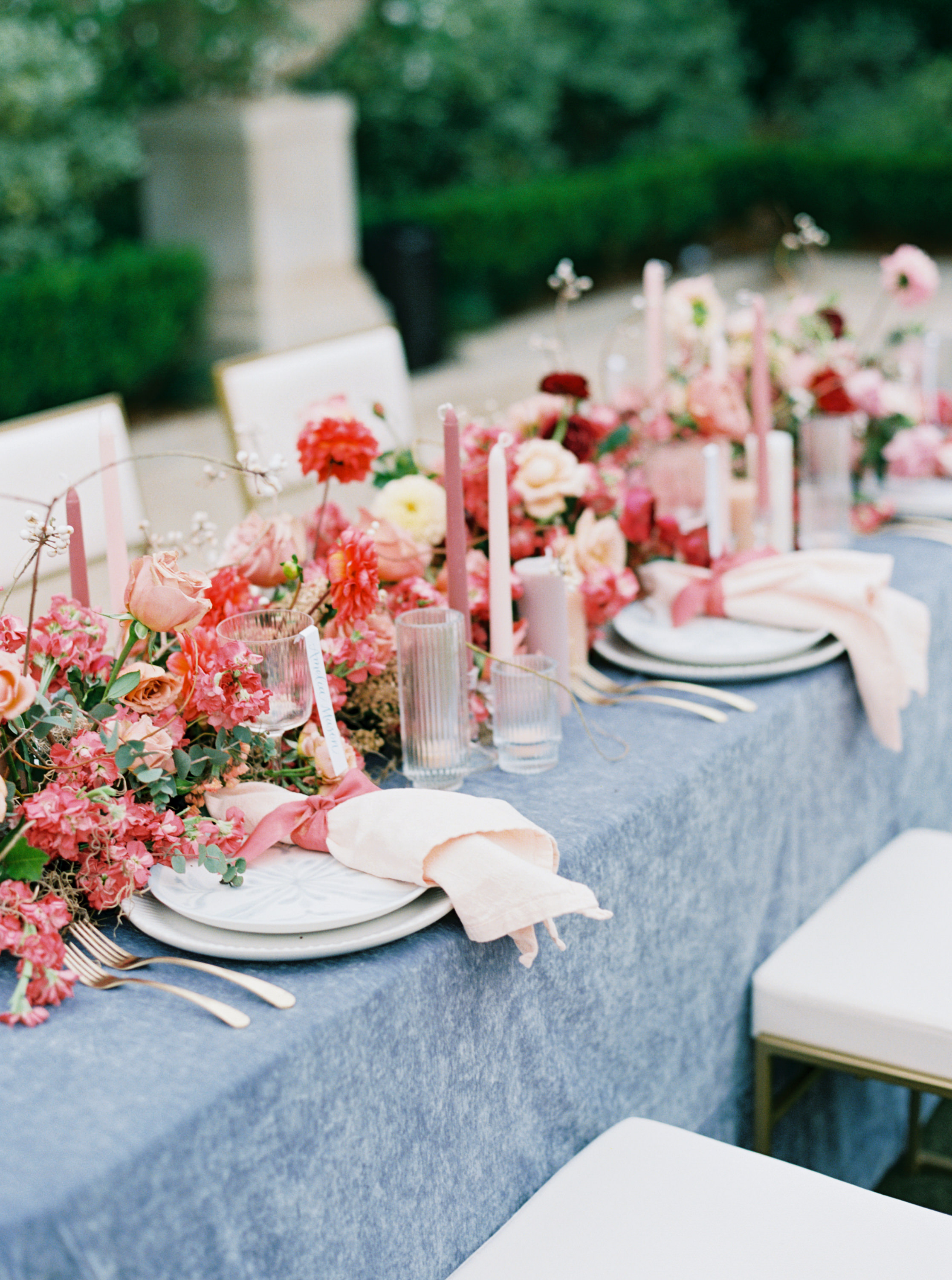 An outdoor wedding table setting with a blue tablecloth, ribbed glassware, and a lush runner of pink, coral, and blush flowers.