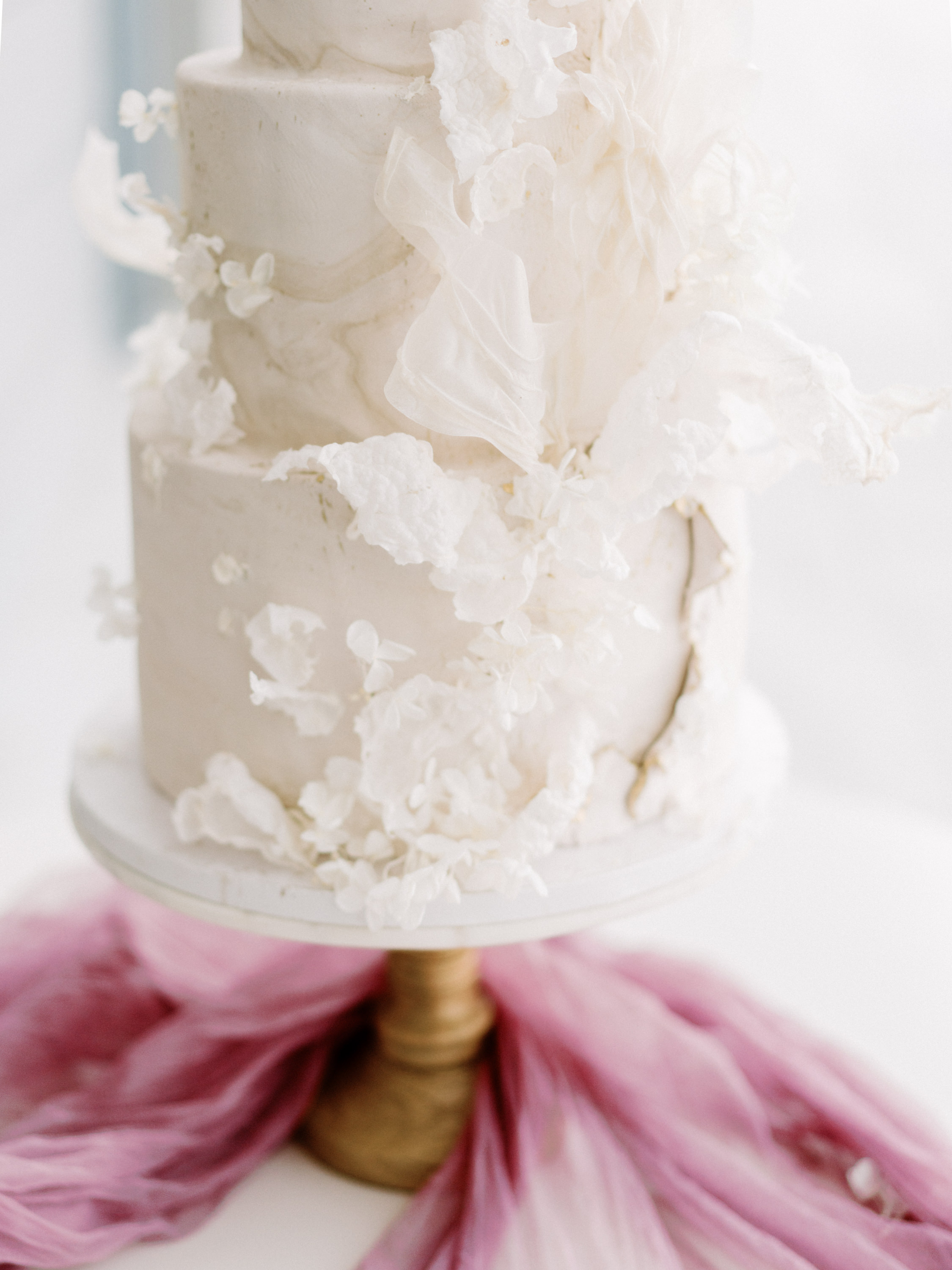 Close-up of a tiered cream wedding cake adorned with delicate ruffled sugar florals and textures on a cake stand.