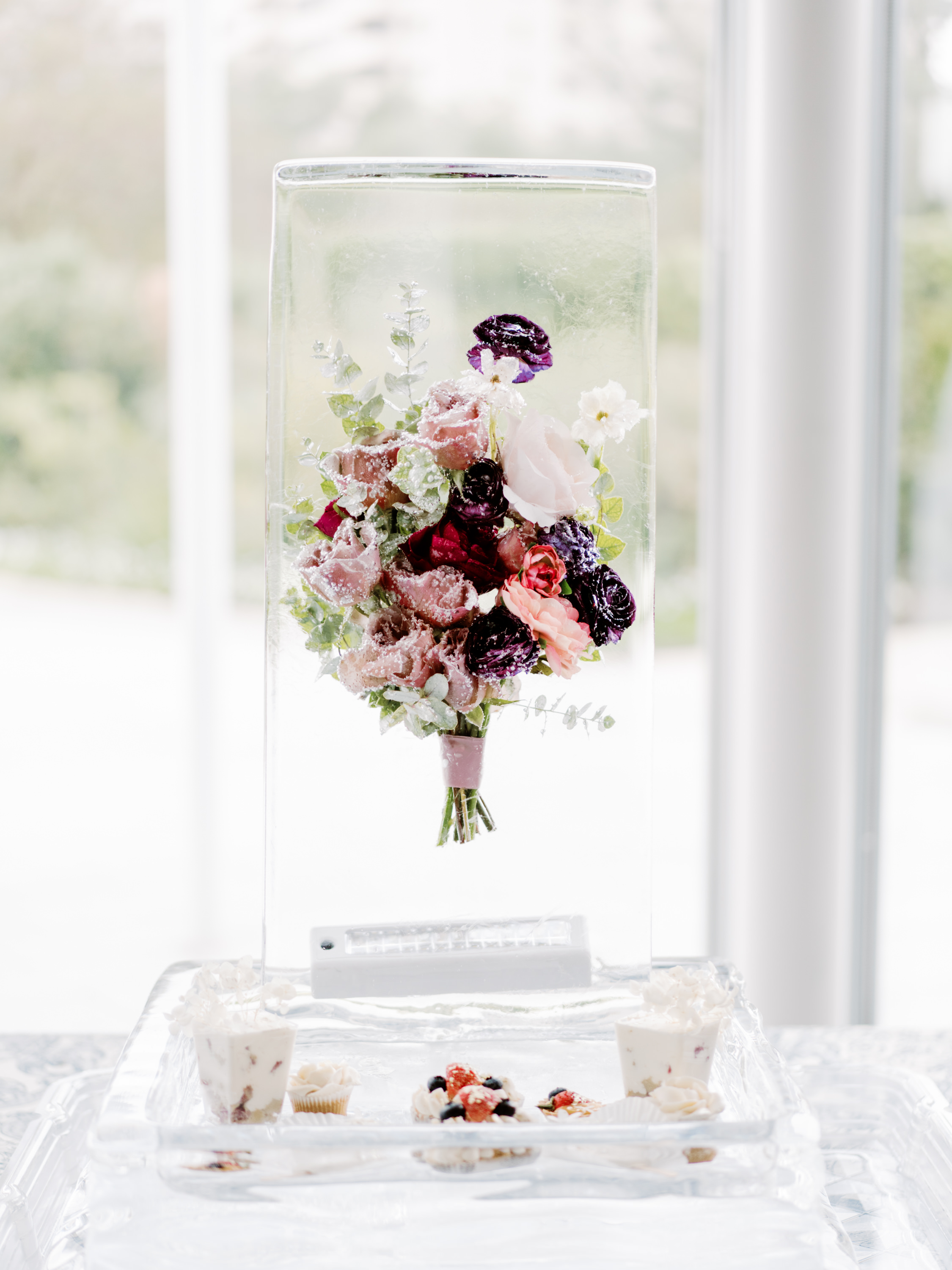 A floral bouquet in shades of dusty rose, plum, and blush frozen inside a tall, clear block of ice.