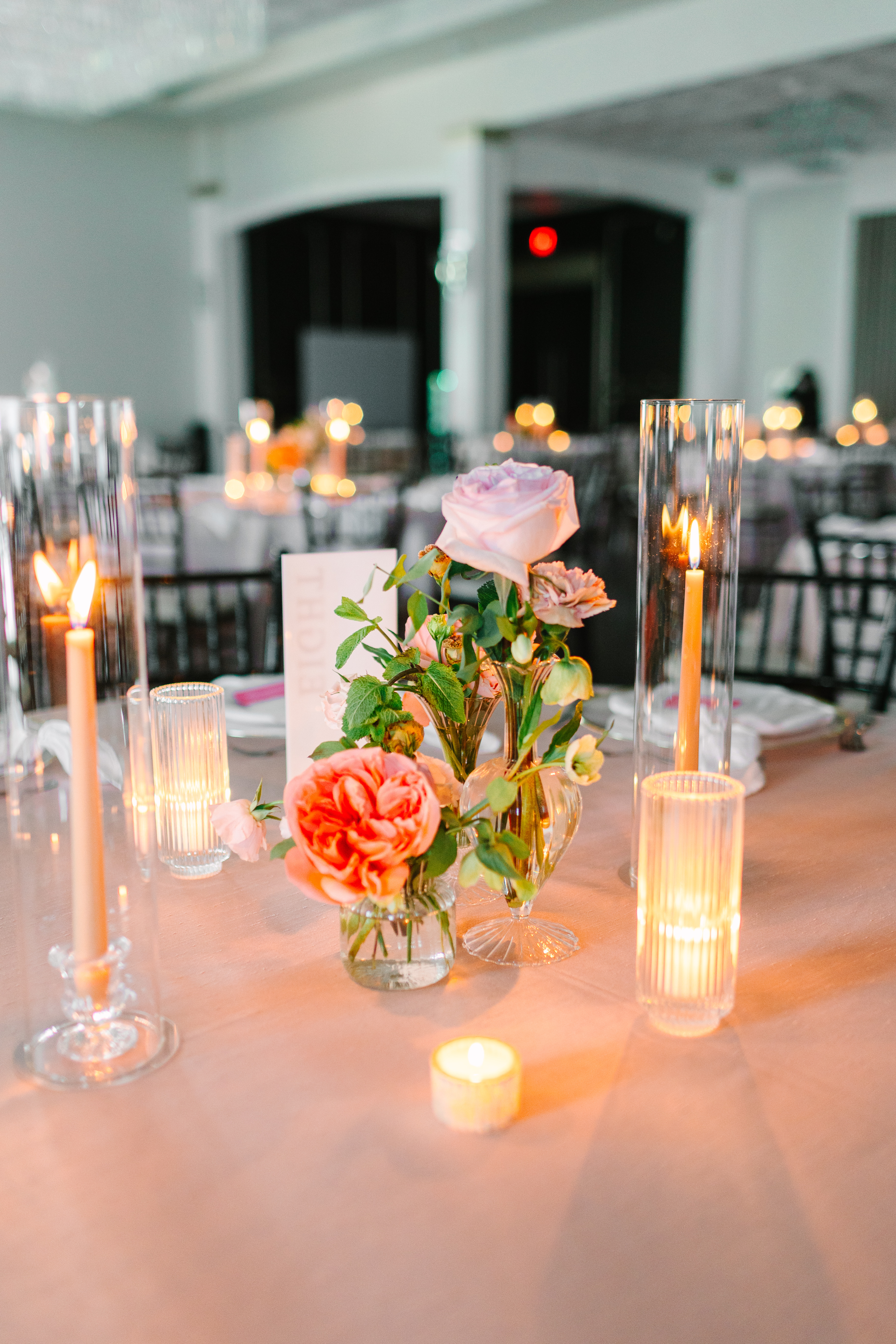 Blush and peach roses in clear glass bud vases beside a glowing votive candle on an evening reception table.