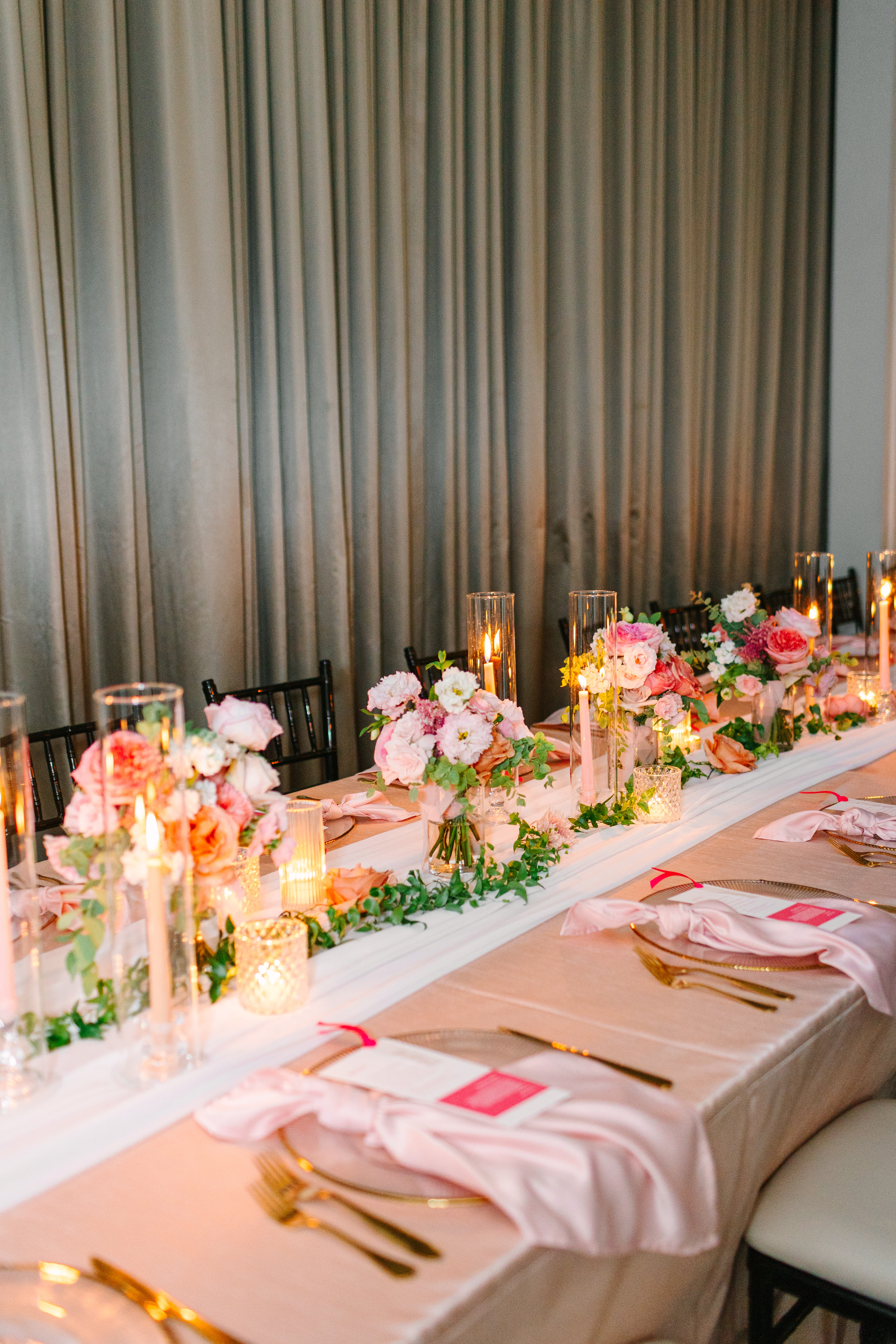 A romantic reception table set with blush pink florals, lit taper candles in glass cylinders, and draped fabric backdrop.