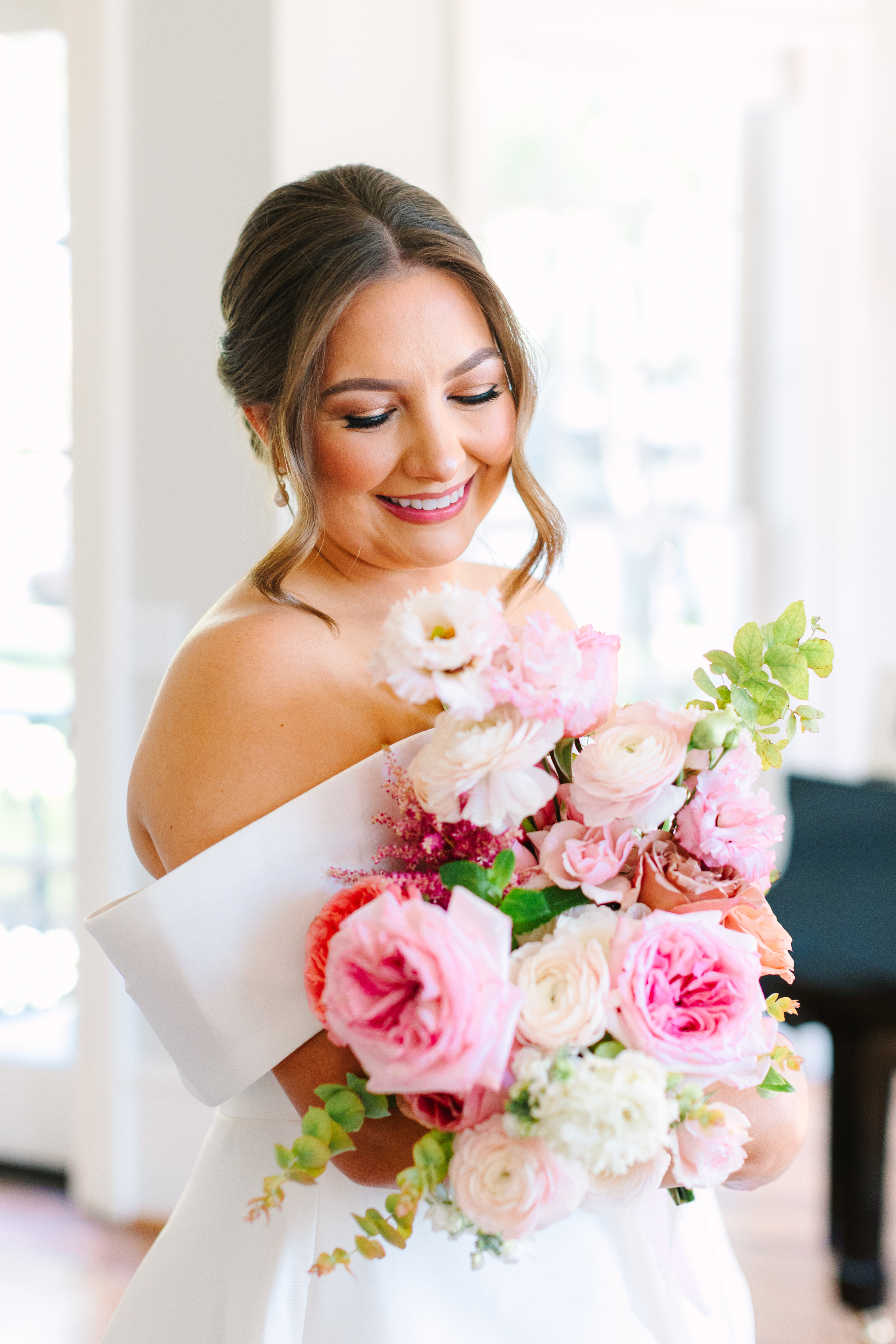 A smiling bride gazes down at a lush bouquet of pink garden roses, blush ranunculus, and green foliage.