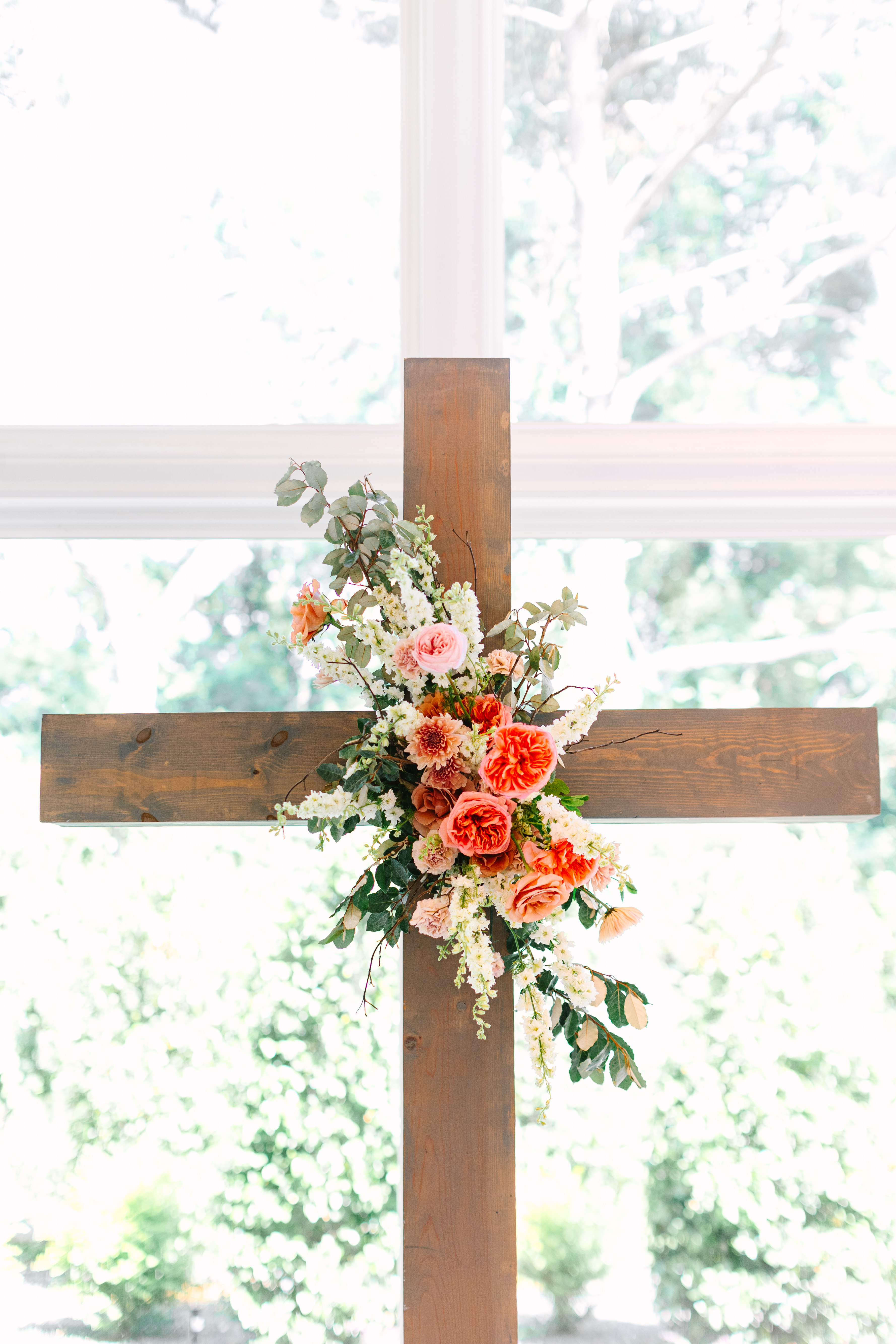 Peach and coral roses with white florals and greenery decorate a wooden ceremonial cross set before a bright window.