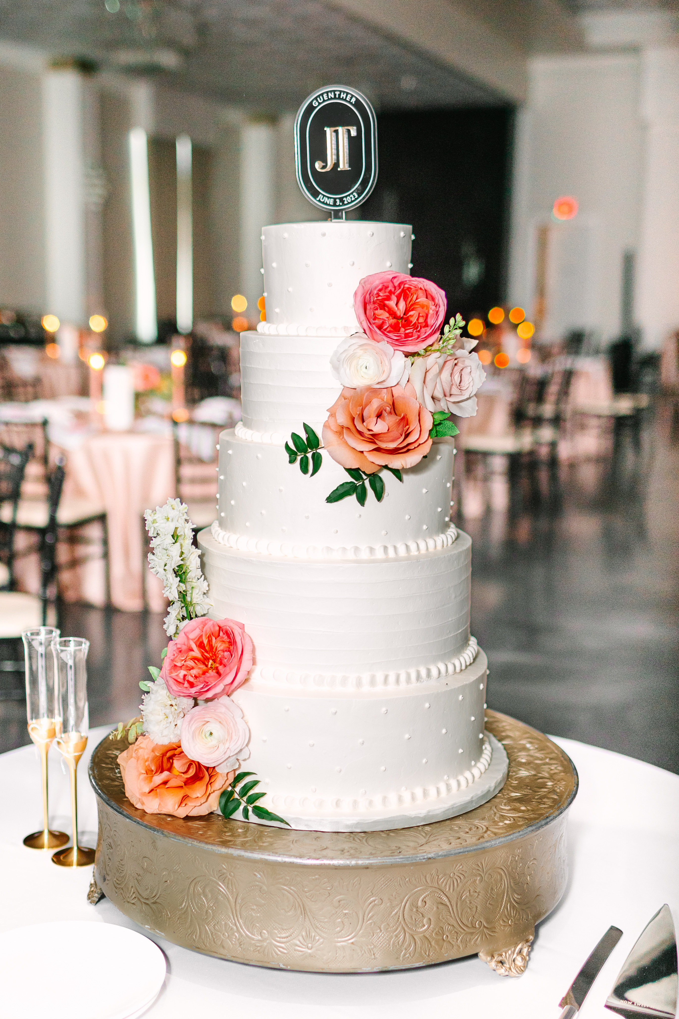 A tiered white wedding cake decorated with fresh coral, peach, and blush roses on an ornate metallic stand.