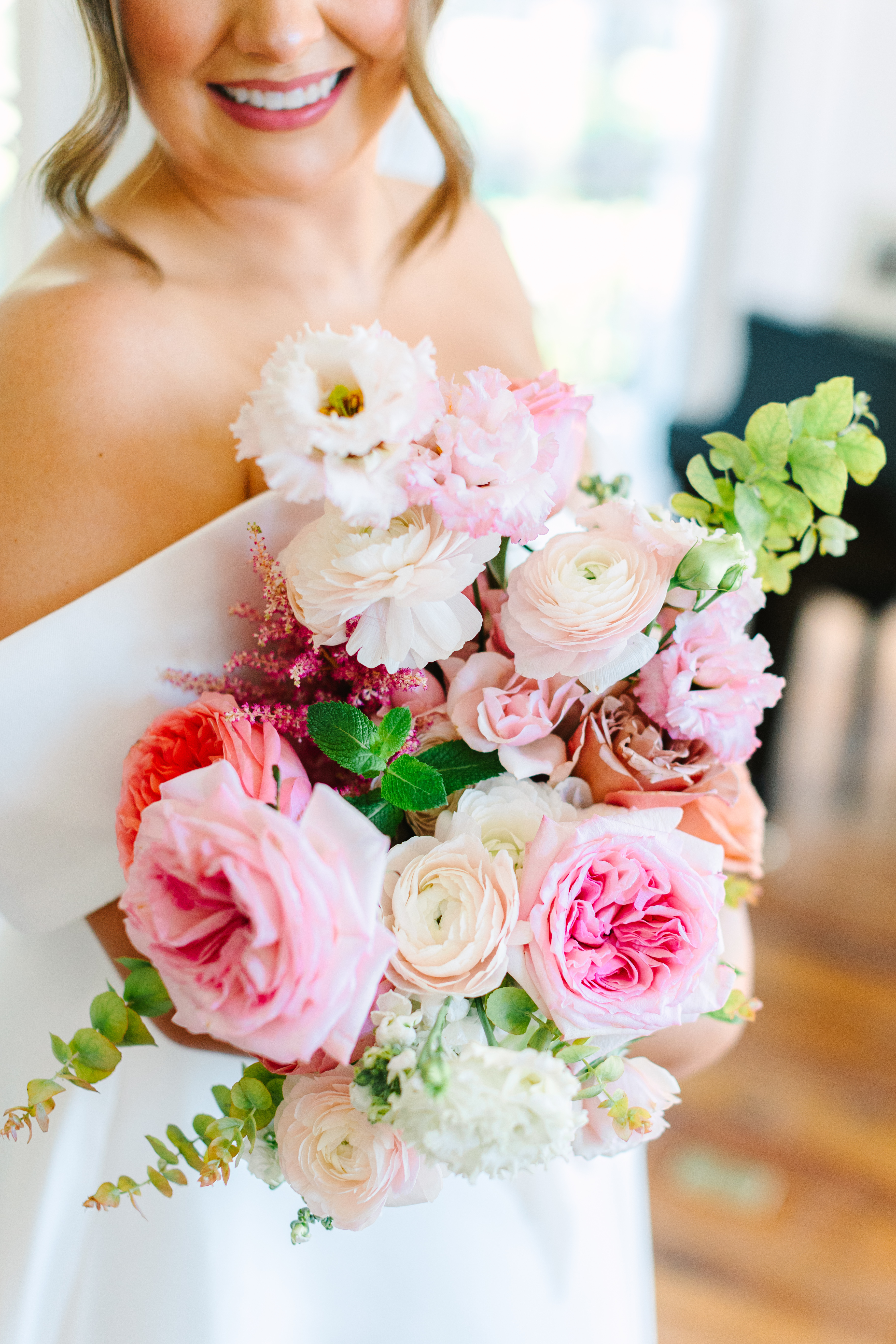 A smiling bride holds a lush floral bouquet featuring blush ranunculus, soft pink roses, and lisianthus blooms.