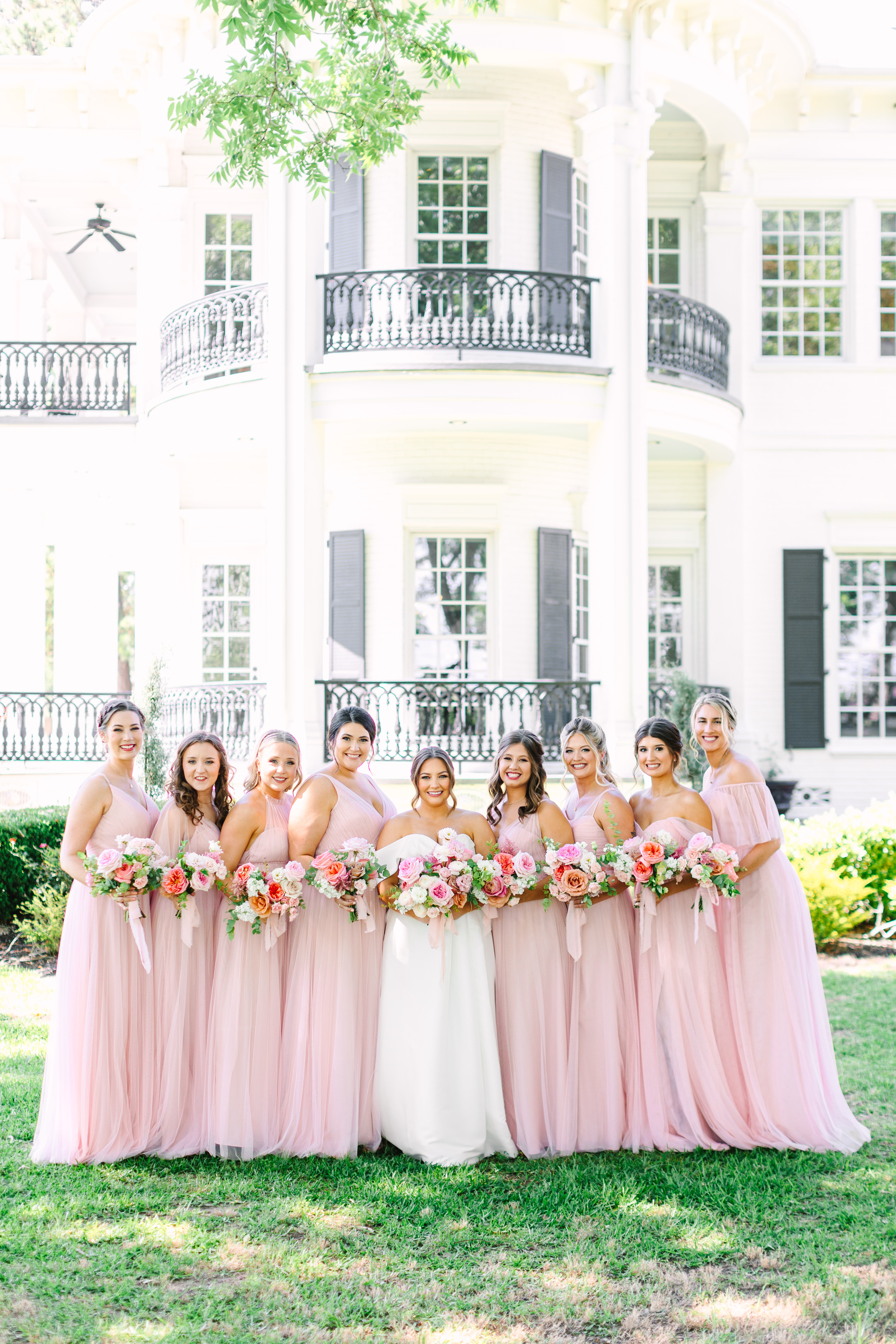 Bride and bridesmaids in blush pink holding pastel rose bouquets in front of an elegant white manor.