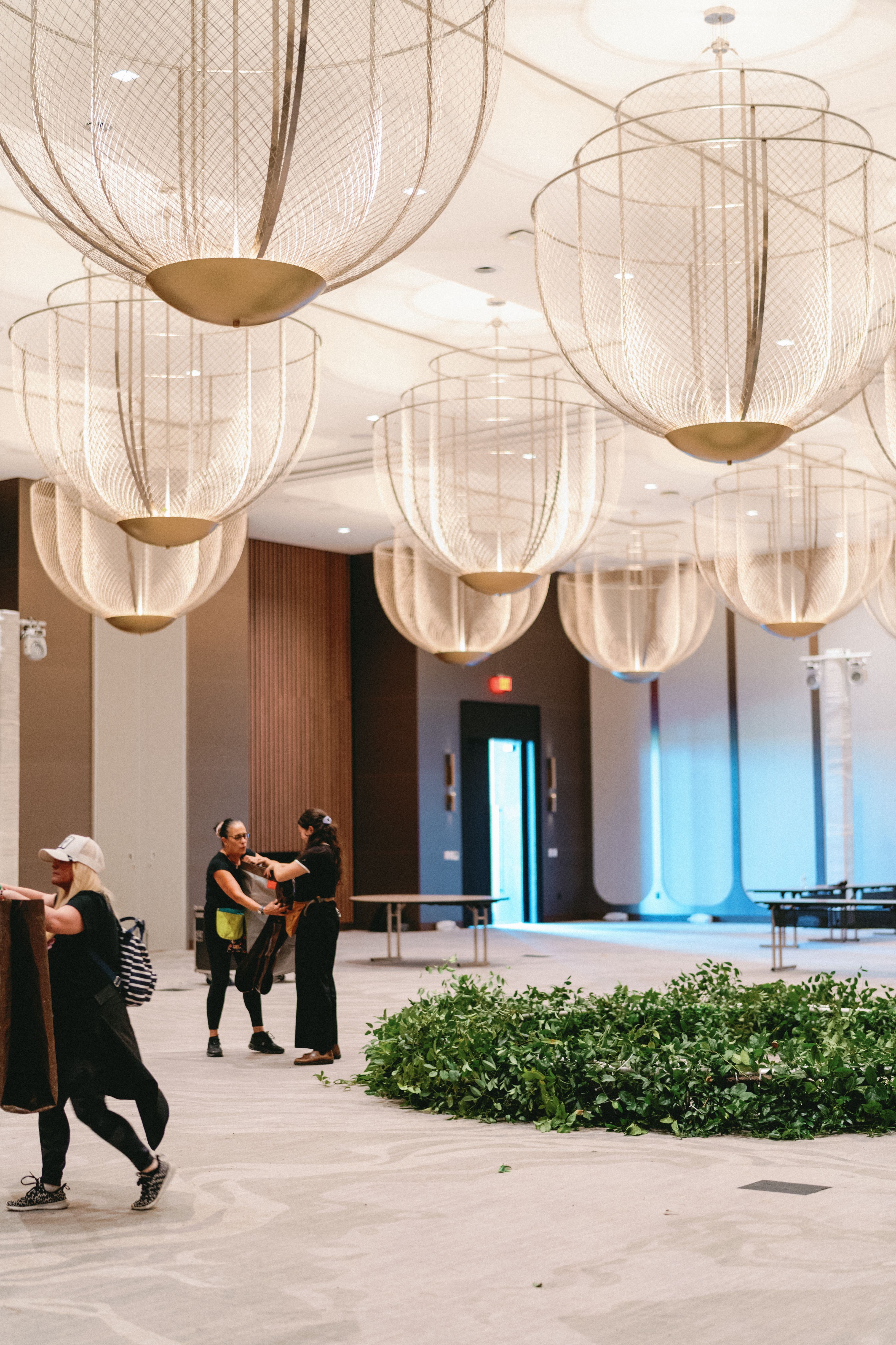 A lush circular greenery arrangement on a carpeted ballroom floor beneath sculptural wire mesh chandeliers.