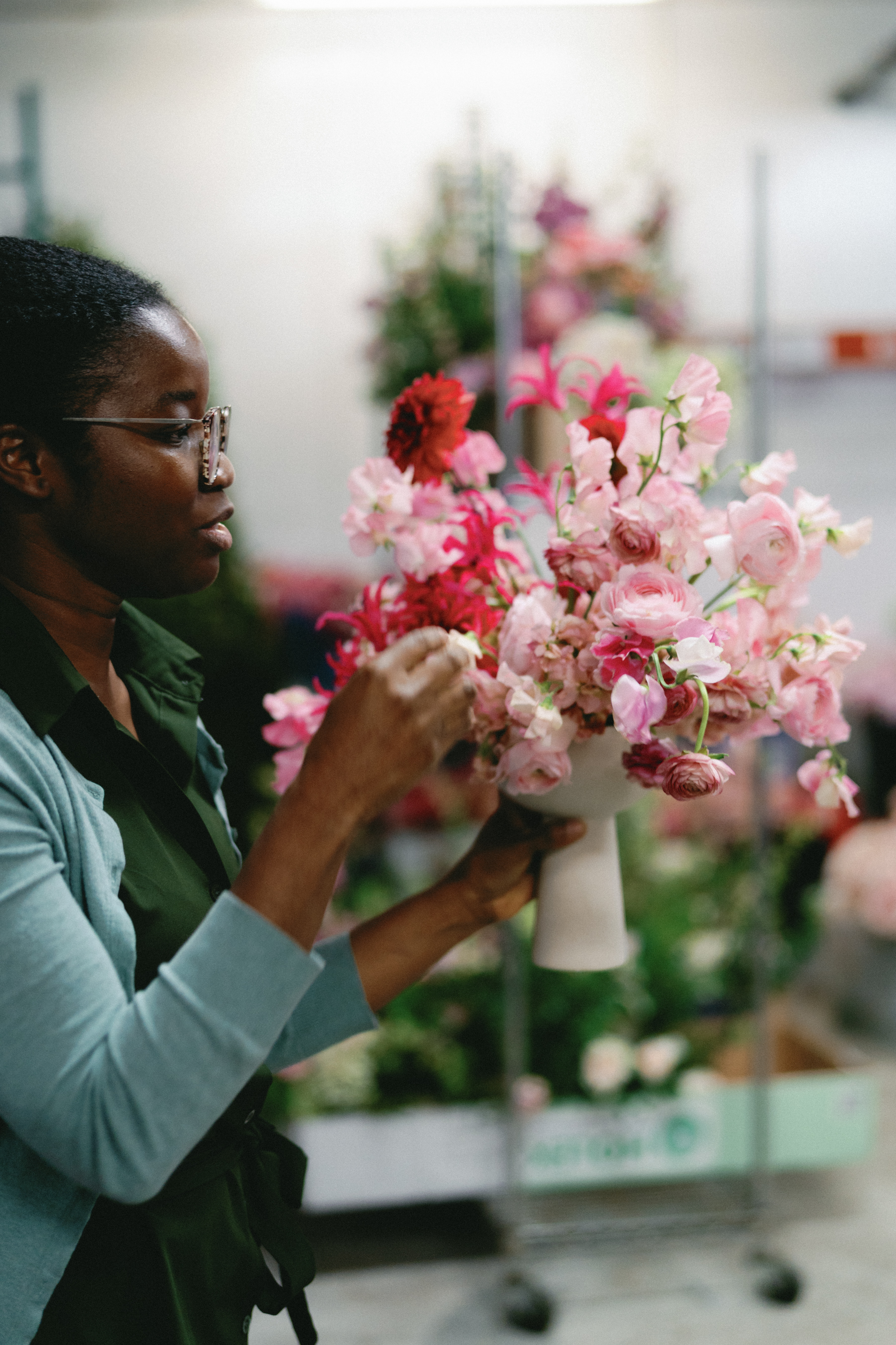 Hands arrange a lush floral centerpiece of light pink sweet peas, bright magenta nerines, and deep red blooms in a white ceramic vase.