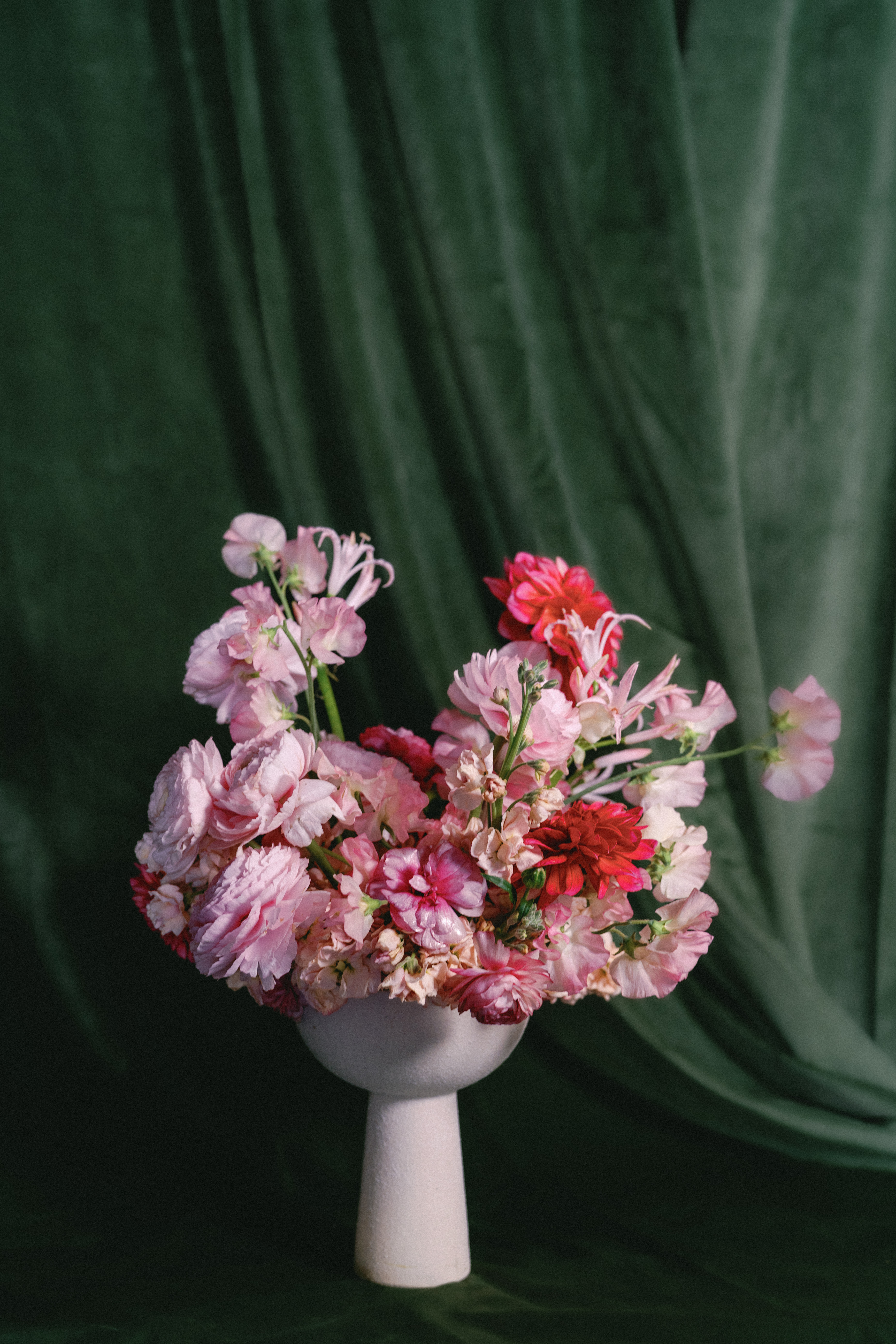 A floral arrangement of pink and vibrant red blossoms sits in a white pedestal vase against a deep green backdrop.