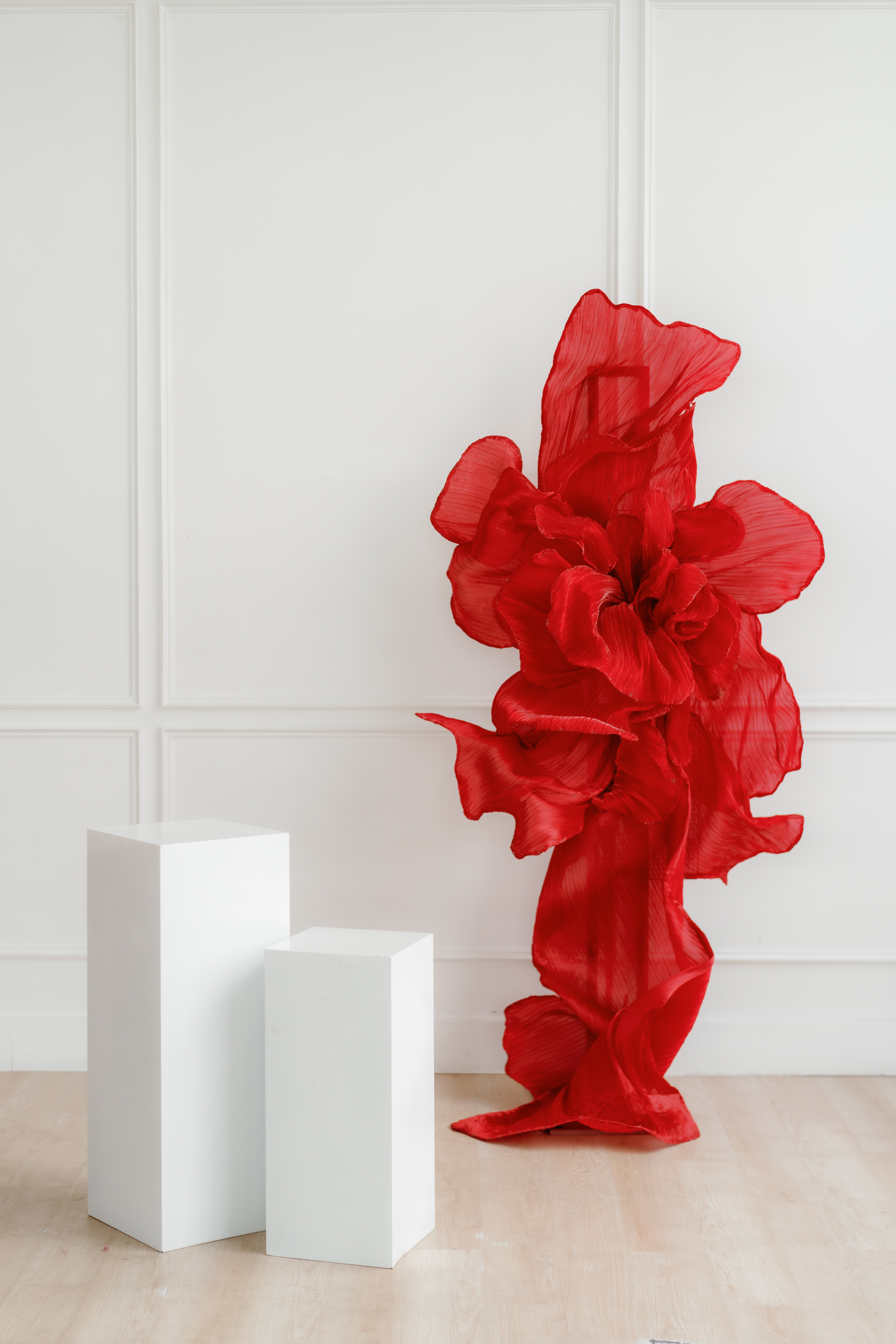 A vibrant red sculptural installation with oversized petal-like forms stands beside a white pedestal in a minimalist room.