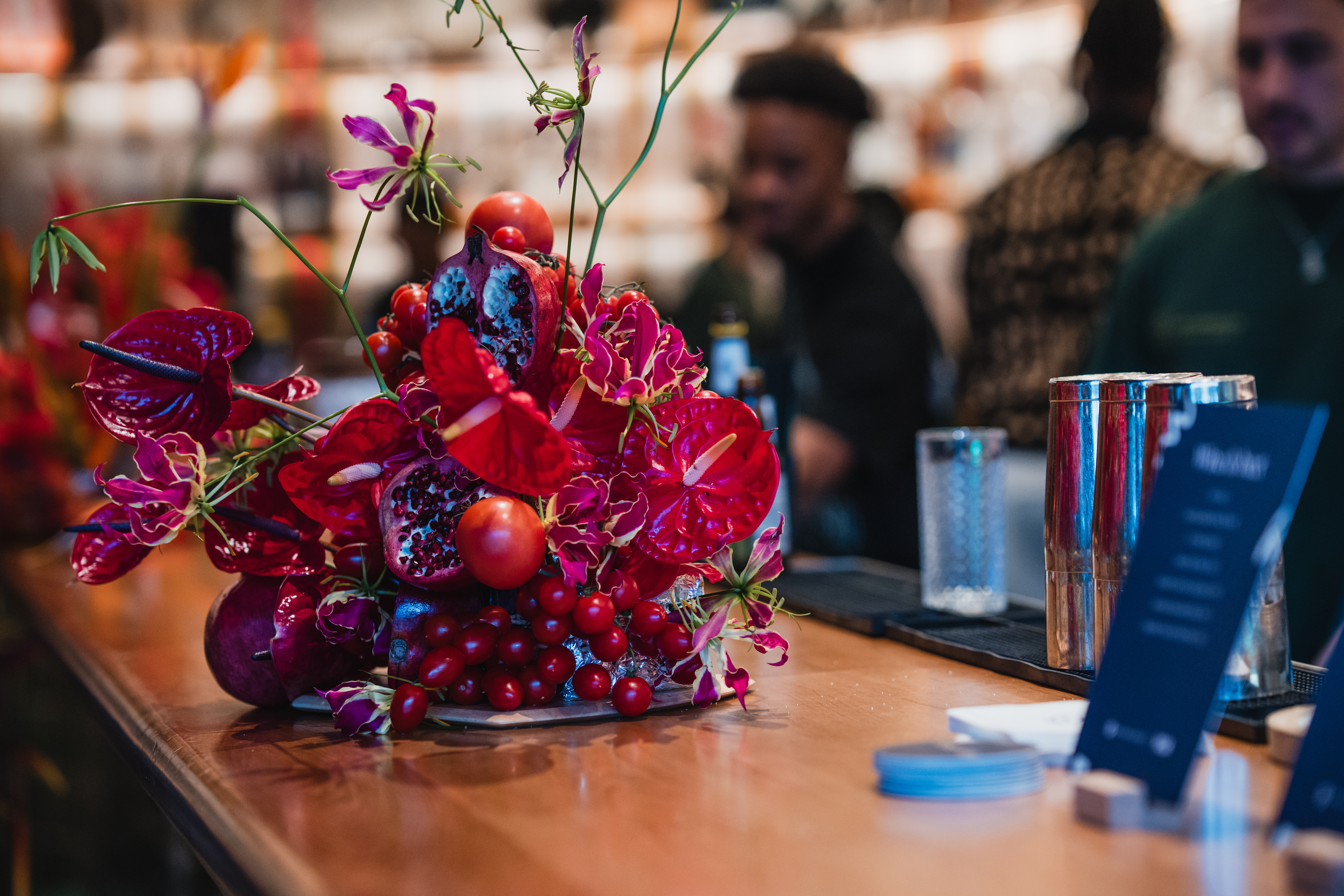 A vibrant tabletop floral arrangement features glossy red anthurium, magenta gloriosa lilies, and cherry tomatoes on a wooden table.