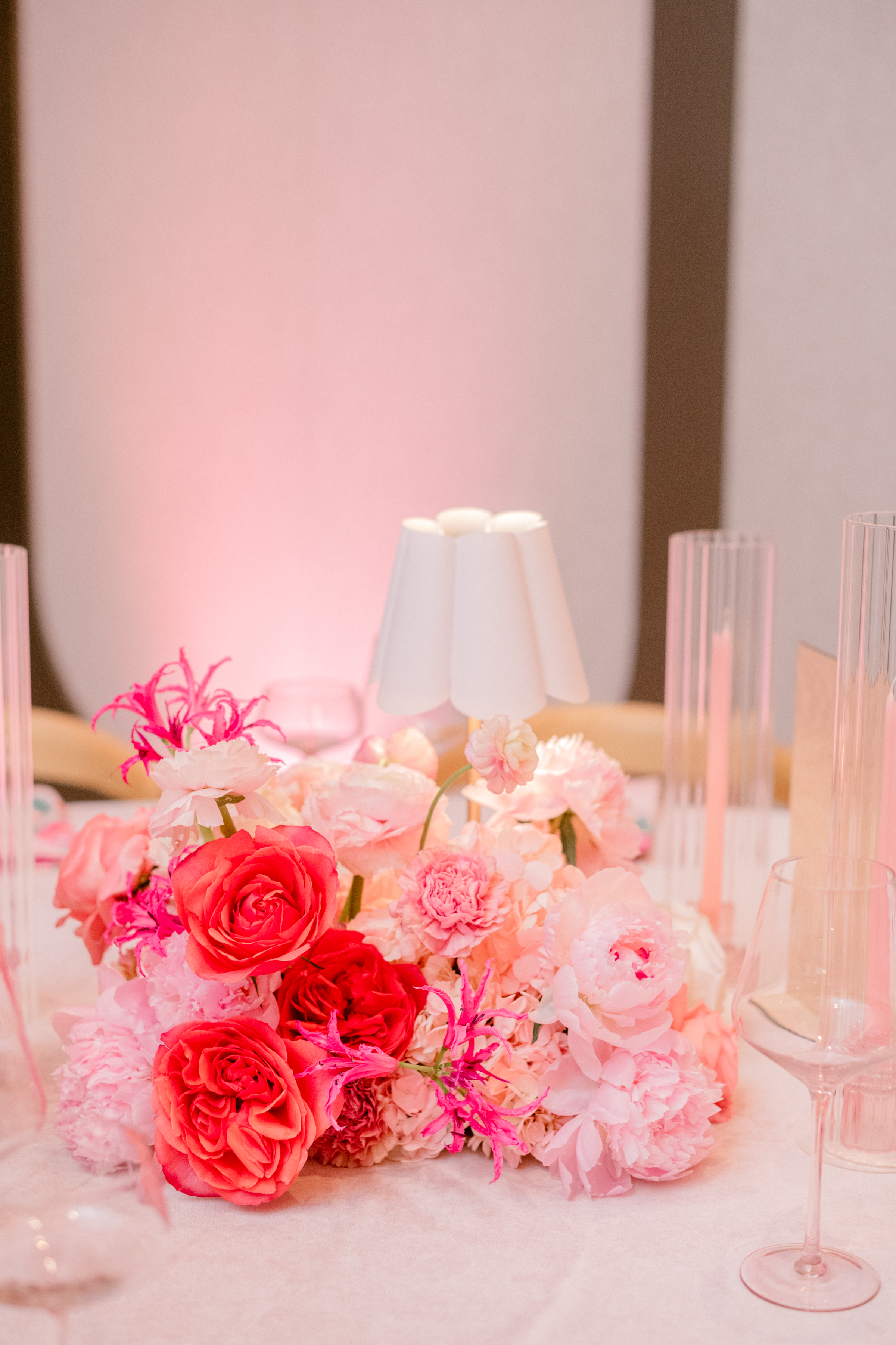 Lush pink peonies, vibrant roses, and a small scalloped table lamp glow in soft ambient lighting on a celebration table.
