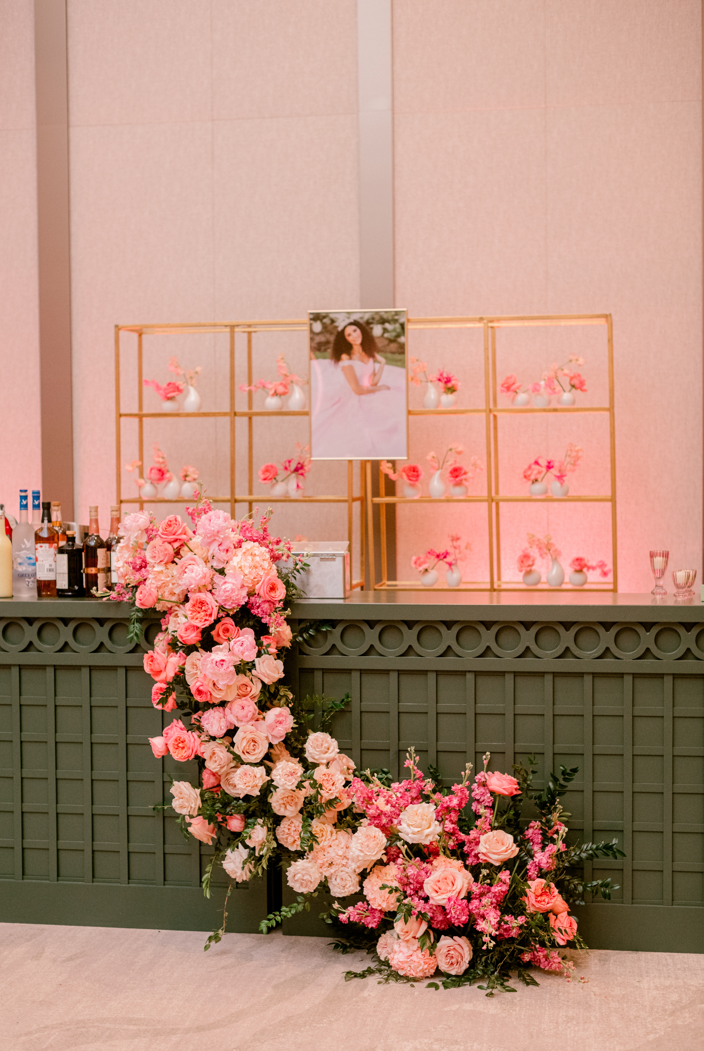 Lush pink and blush floral garland cascades down an olive green latticed bar in front of gold shelving displays.