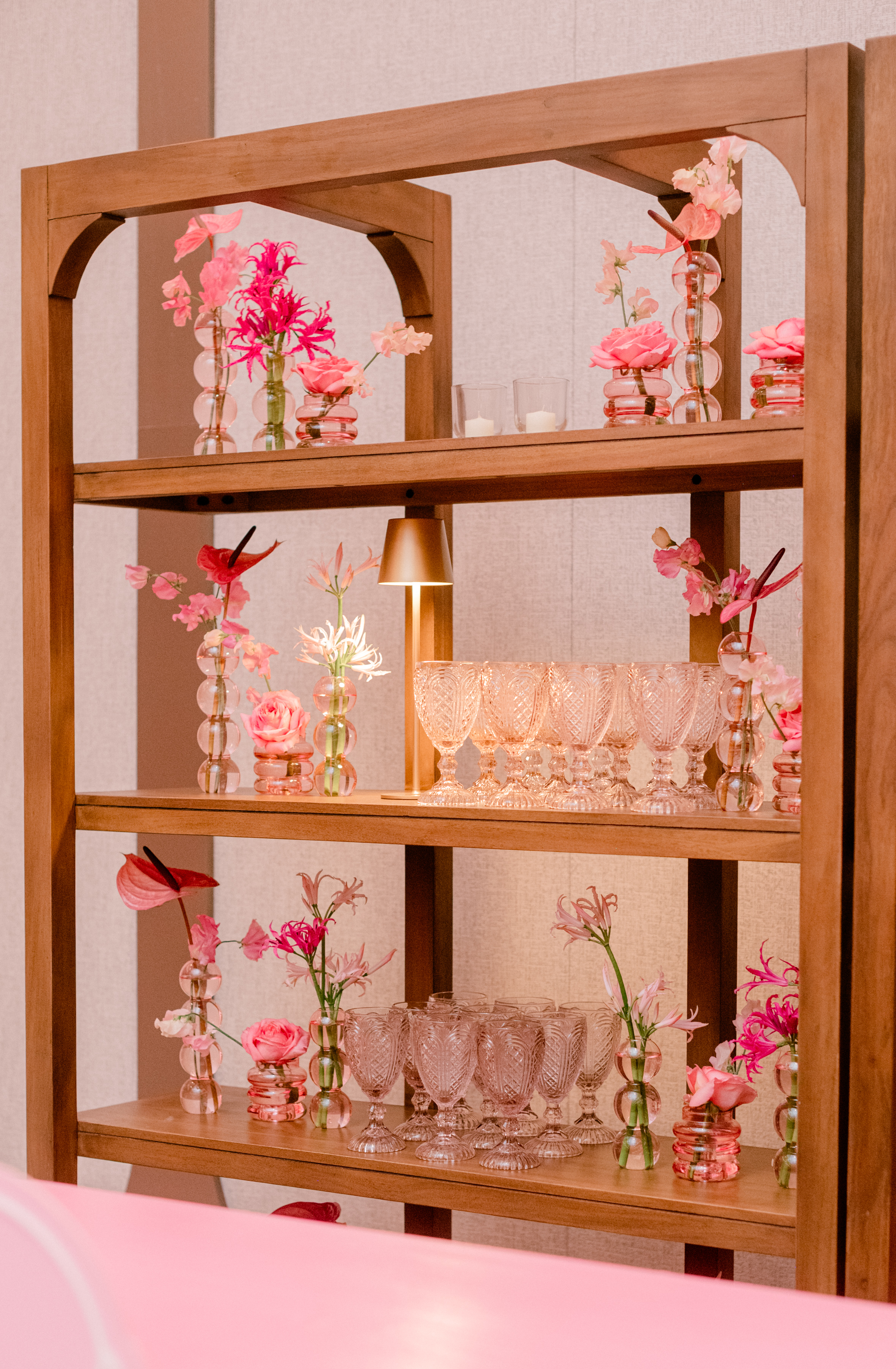Wooden shelving styled with pink cut-glass goblets, small brass lamps, and vibrant pink floral bud vases.