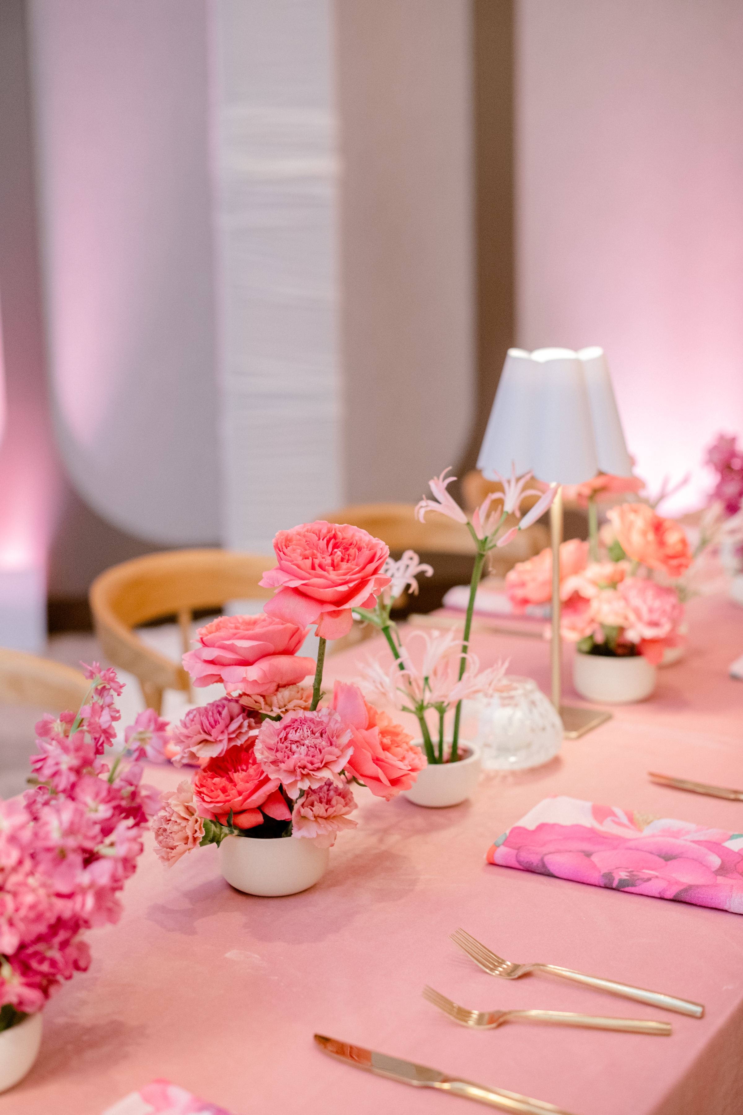 Bright pink roses and carnations in white ceramic bowls arranged on a pink tablecloth with gold flatware.