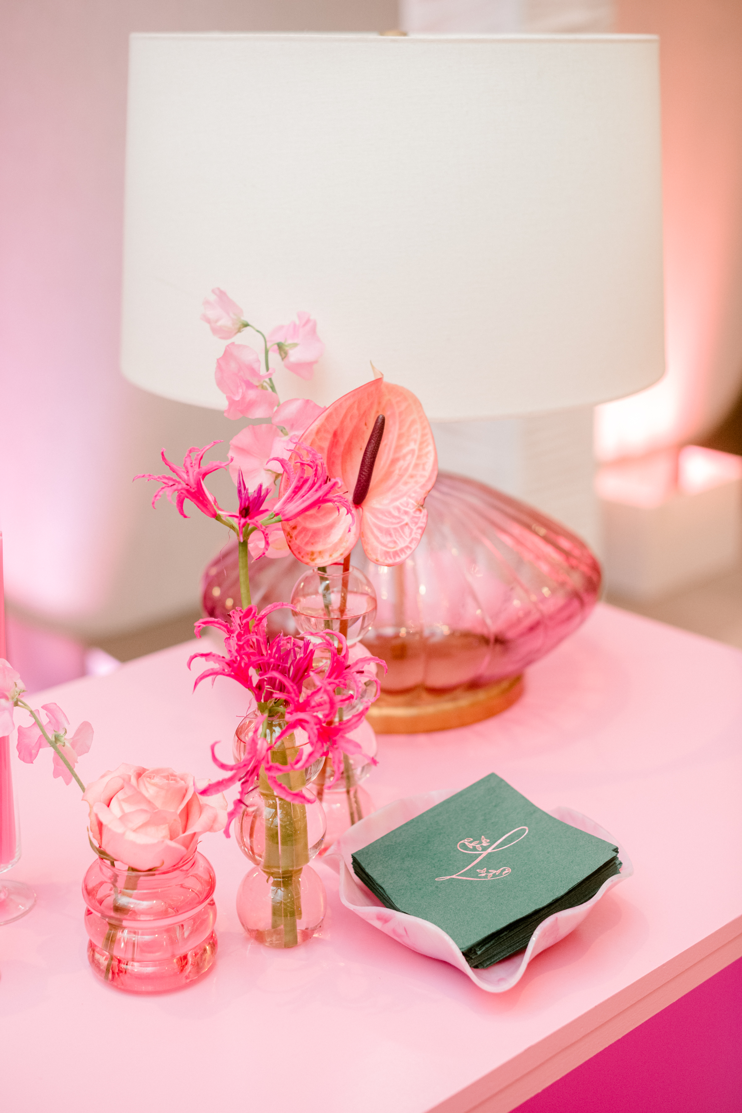Vibrant pink florals in glass bud vases beside monogrammed green napkins and a fluted glass lamp.