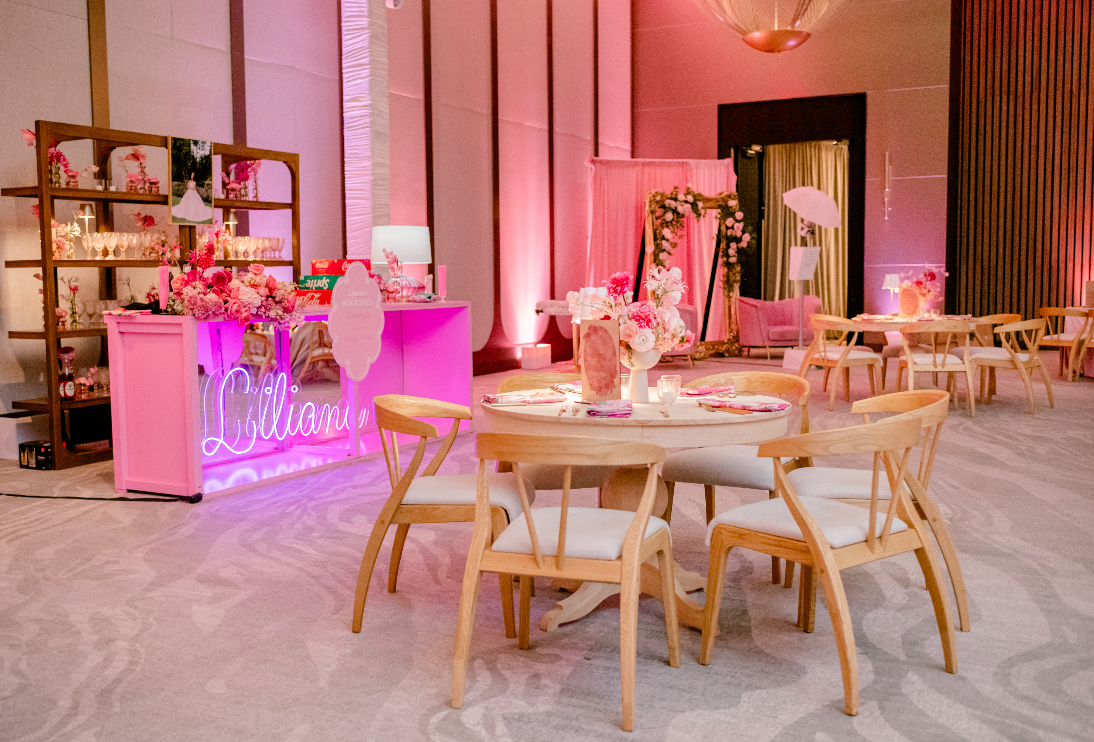 Round wooden dining table with a pink floral centerpiece and mid-century chairs under glowing pink event lighting.