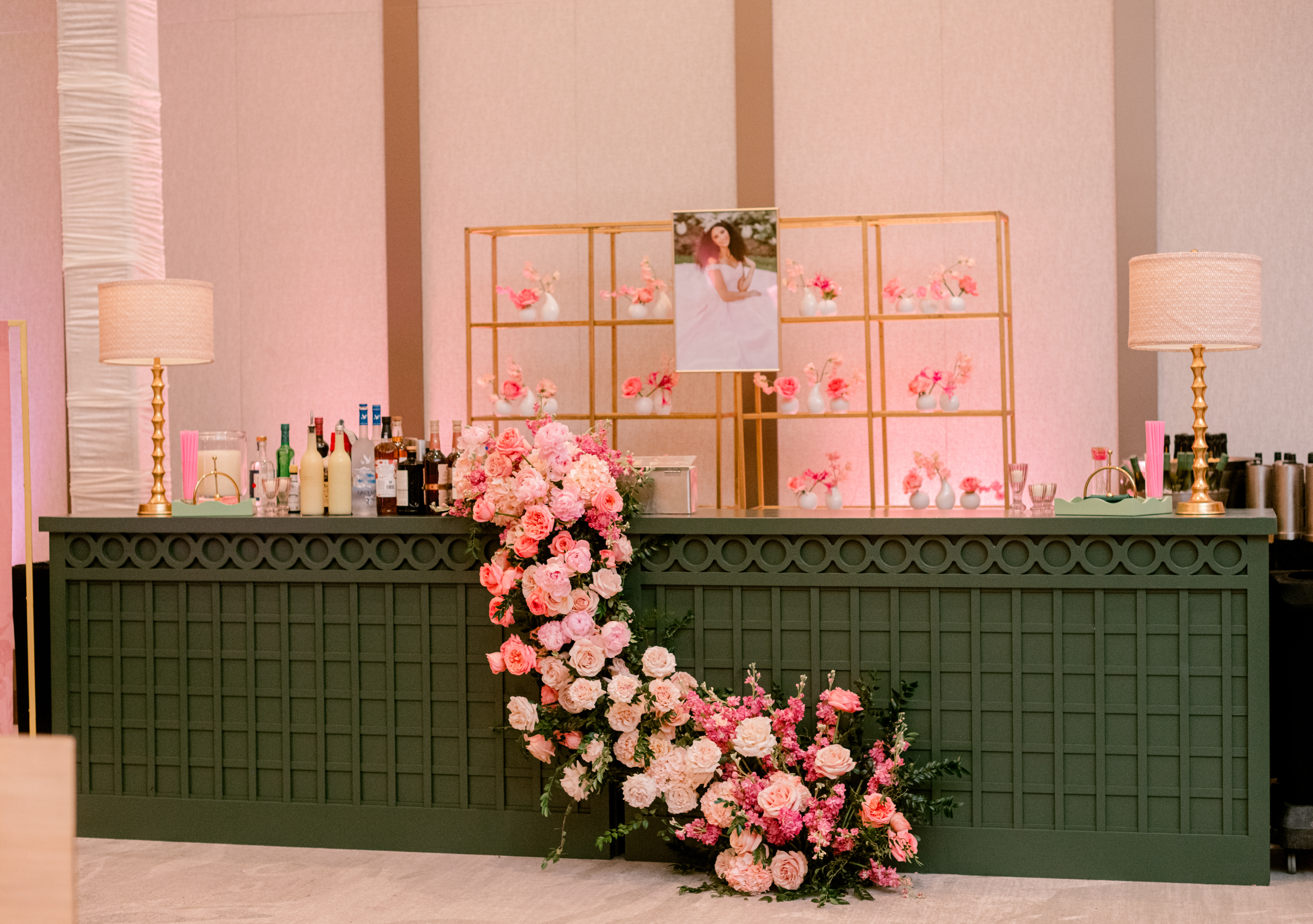 A dark green event bar draped with a cascading floral installation of blush and pink roses in front of gold display shelving.