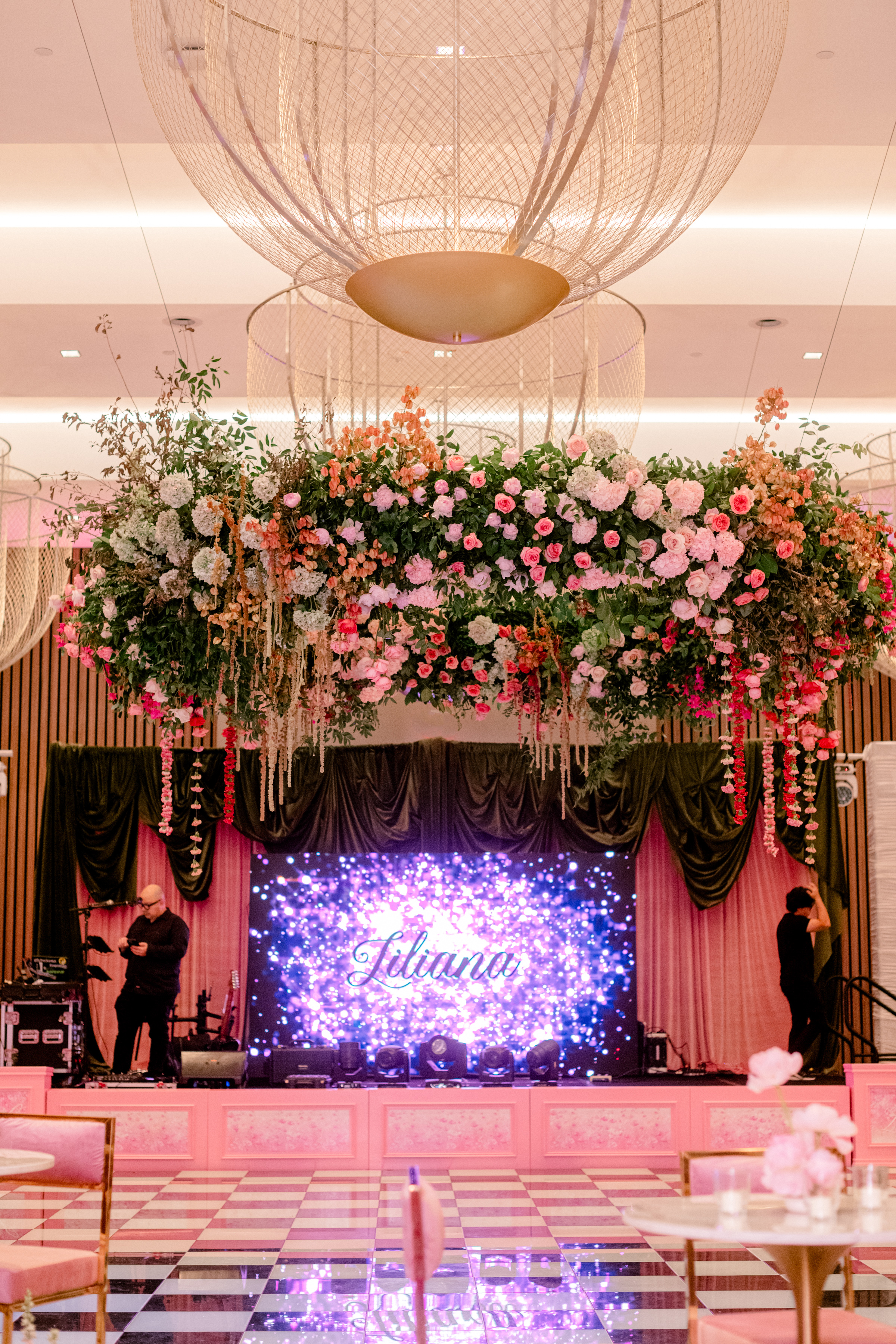 Suspended floral installation of pink roses and greenery beneath a sculptural chandelier overlooking a stage with a checkered dance floor.
